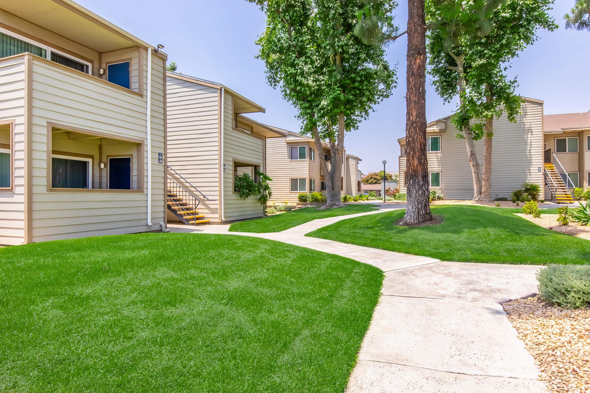 A view of a landscaped area with well-maintained green lawns surrounding modern apartment buildings. The buildings are light-colored with balconies, and there are trees providing shade. A concrete pathway connects the buildings, creating a welcoming atmosphere in a residential community.