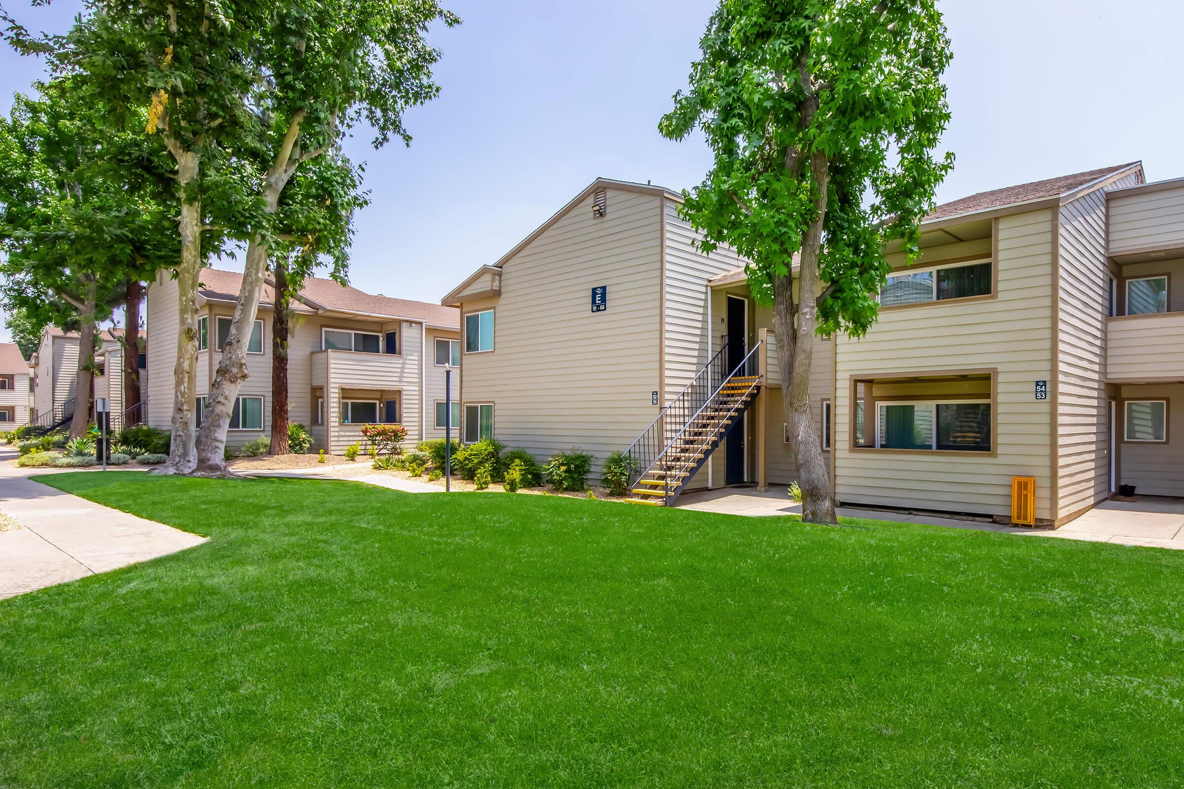 A view of a residential complex featuring two-story apartment buildings with beige siding, surrounded by well-maintained green grass and trees. A pathway leads through the lawn, with a set of stairs leading to the entrance of one of the buildings. The setting appears sunny and inviting.