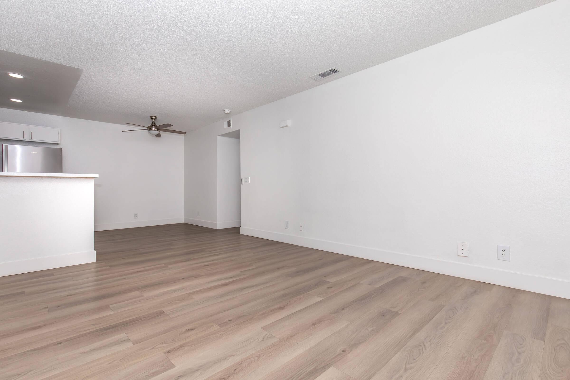 A spacious, empty living area featuring light wood flooring and white walls. There is a ceiling fan installed, and the room includes a kitchen area with a counter. The space is well-lit and has a clean, modern aesthetic.