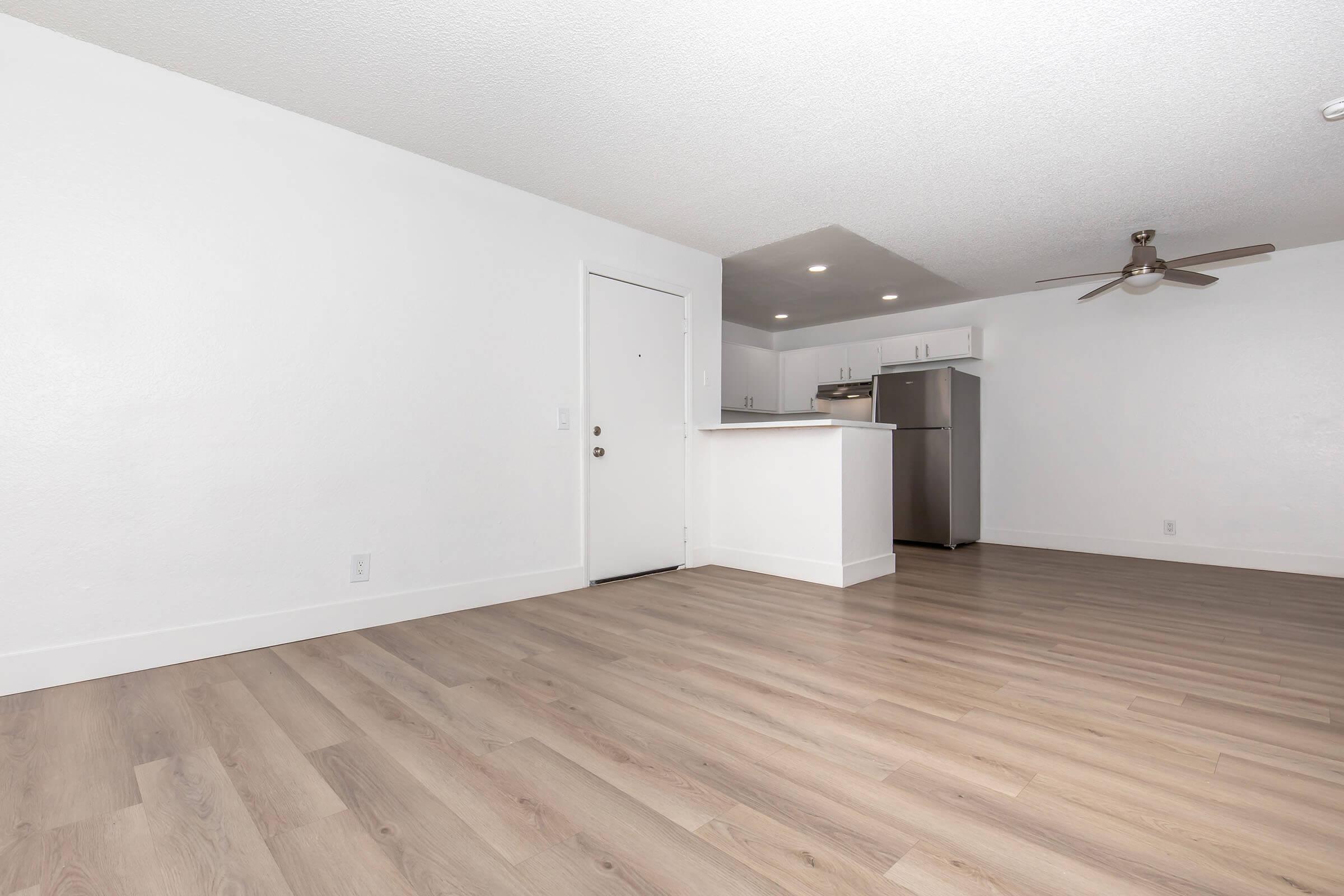 A spacious, modern living area with light-colored walls and hardwood flooring. A white kitchen countertop is visible in the background, along with stainless steel appliances. The entrance door is on the left, and a ceiling fan is installed. The room is well-lit, creating a bright and airy atmosphere.