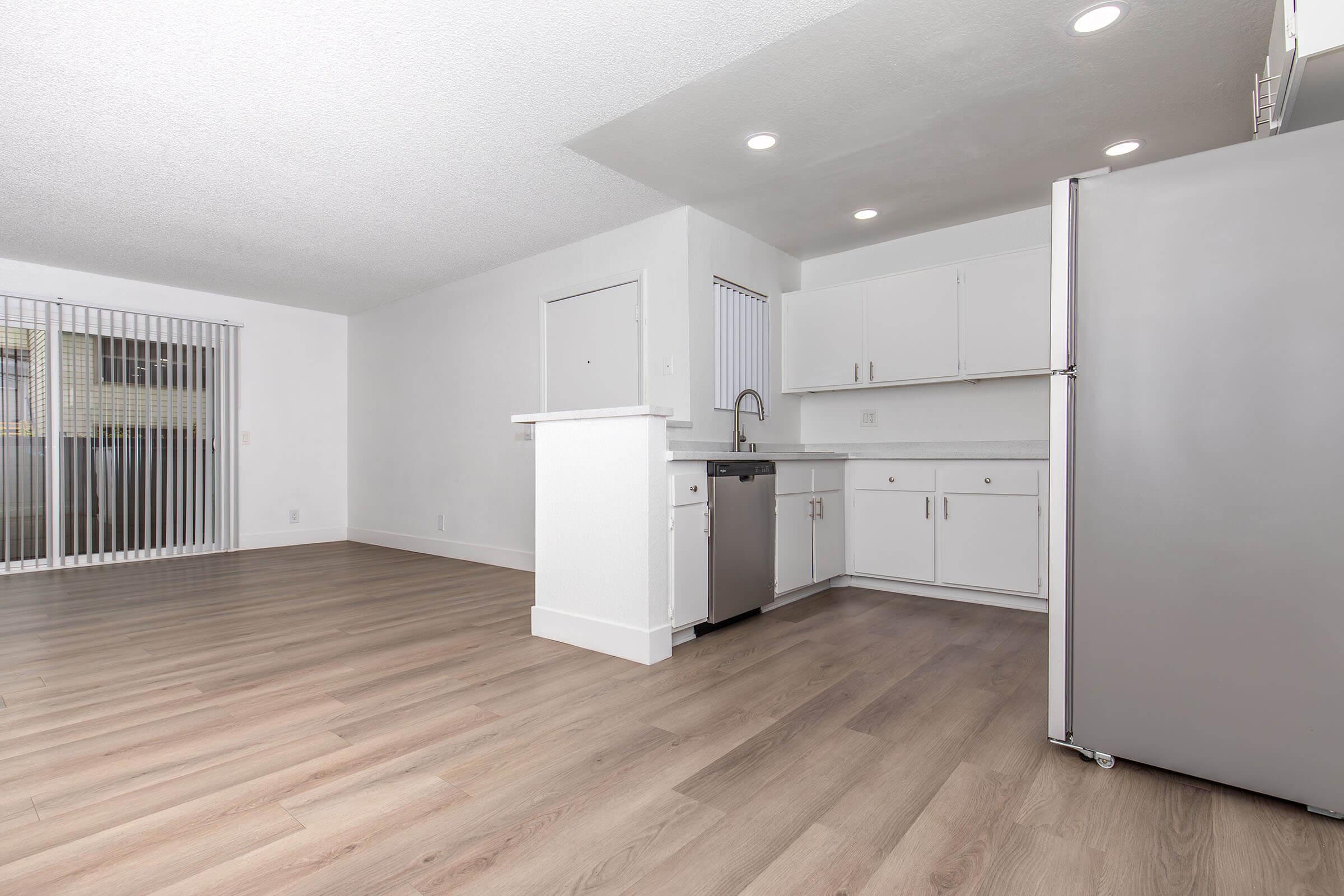 Interior view of a modern kitchen and living area featuring light wood flooring, white cabinetry, and stainless steel appliances. A sliding glass door leads to a patio. The space is well-lit with recessed lighting and has a clean, minimalist aesthetic.