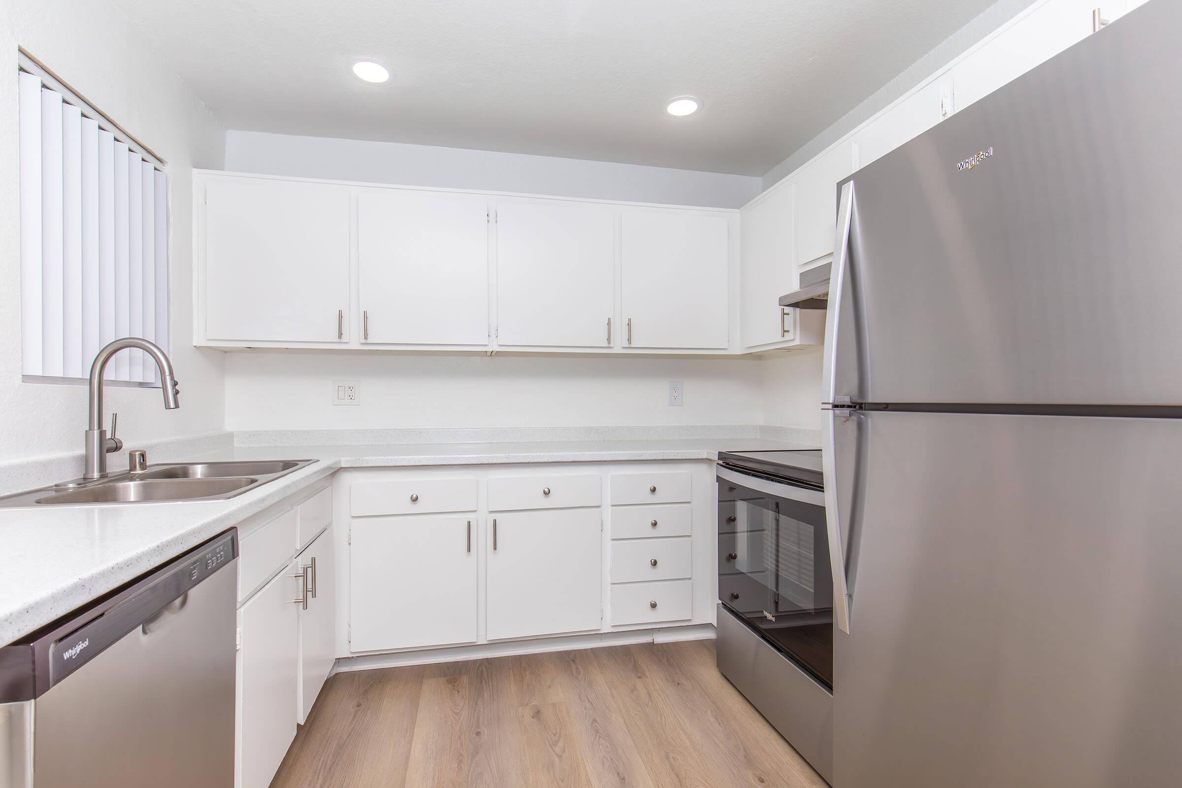 A modern kitchen featuring white cabinets, a stainless steel refrigerator, a black oven, and a sink with a pull-down faucet. The countertops are light-colored, and there is a window with blinds on the left. The flooring is a light wood laminate, creating a clean and bright aesthetic.