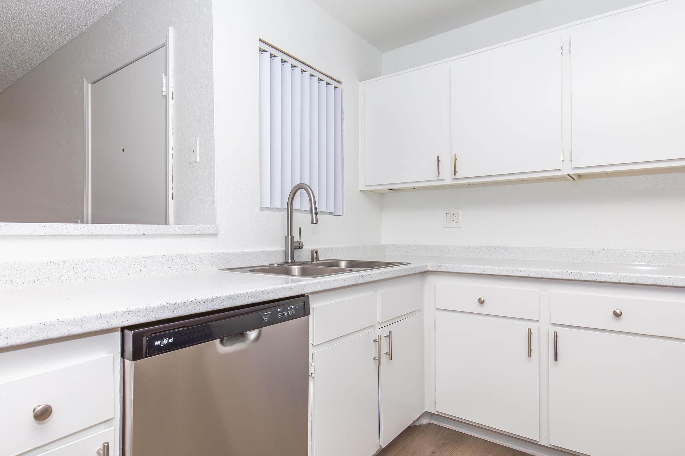 Modern kitchen with white cabinetry and quartz countertops. A stainless steel sink with a sleek faucet is positioned beside a built-in dishwasher. Natural light enters through a window with vertical blinds. The space is bright and minimalistic, featuring simple, contemporary design elements.