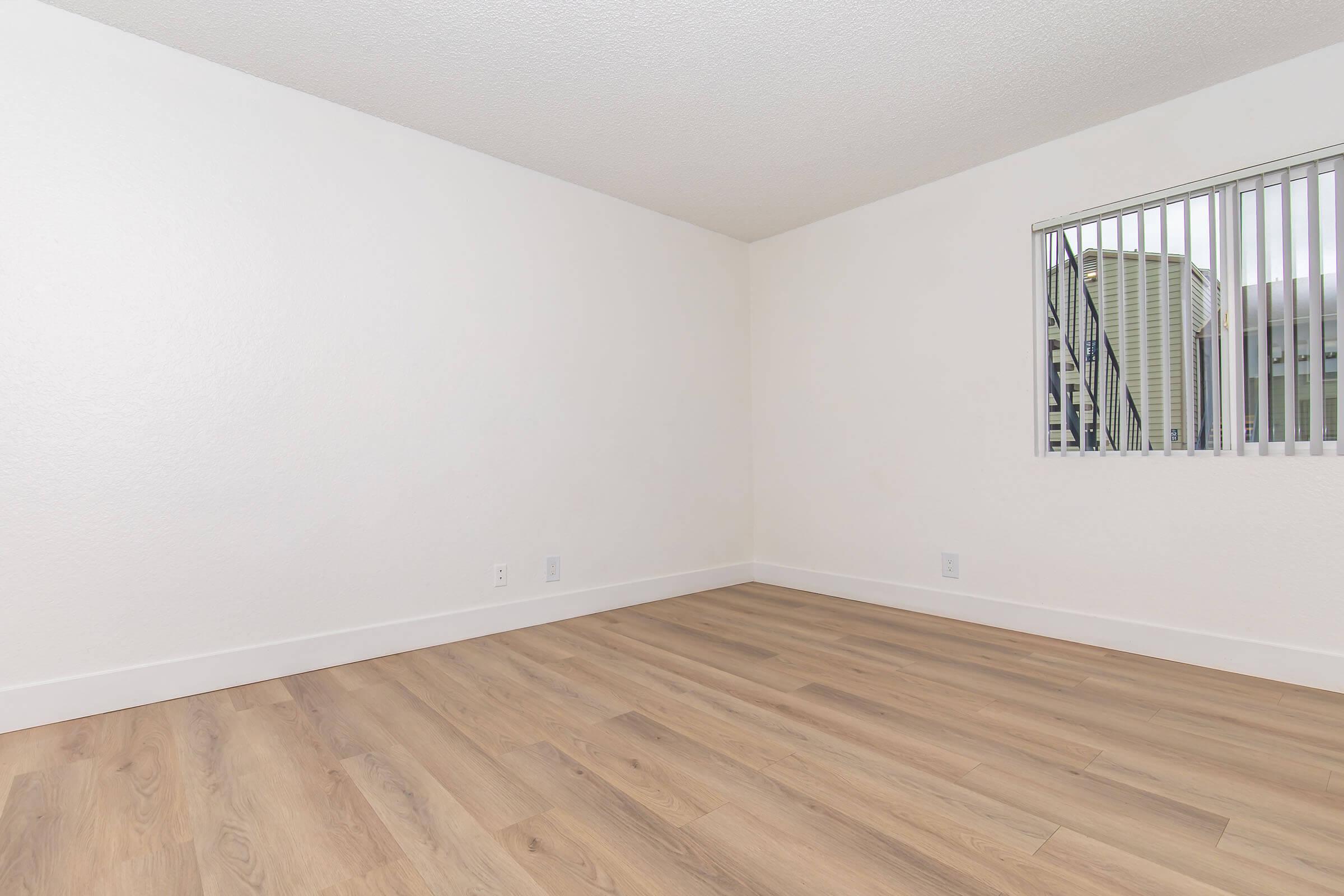 A clean, empty room with light-colored walls and wood-style laminate flooring. There is a window with vertical blinds allowing natural light, and the space appears spacious and well-lit, suitable for various uses. The overall atmosphere is simple and minimalist.