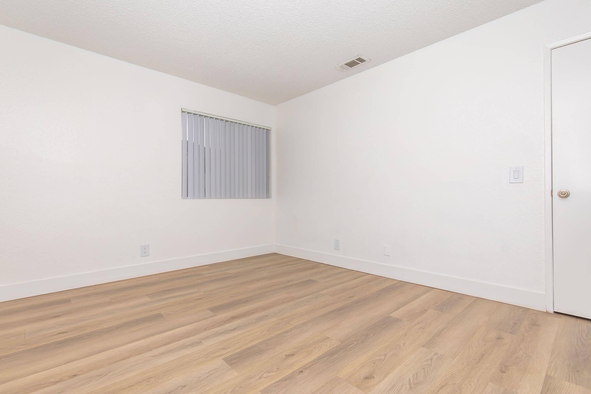 A small, empty room with light wood flooring and plain white walls. A window with vertical blinds is on one wall, and a closed door is visible on another wall. The overall feel is minimalist and bright, with ample natural light.