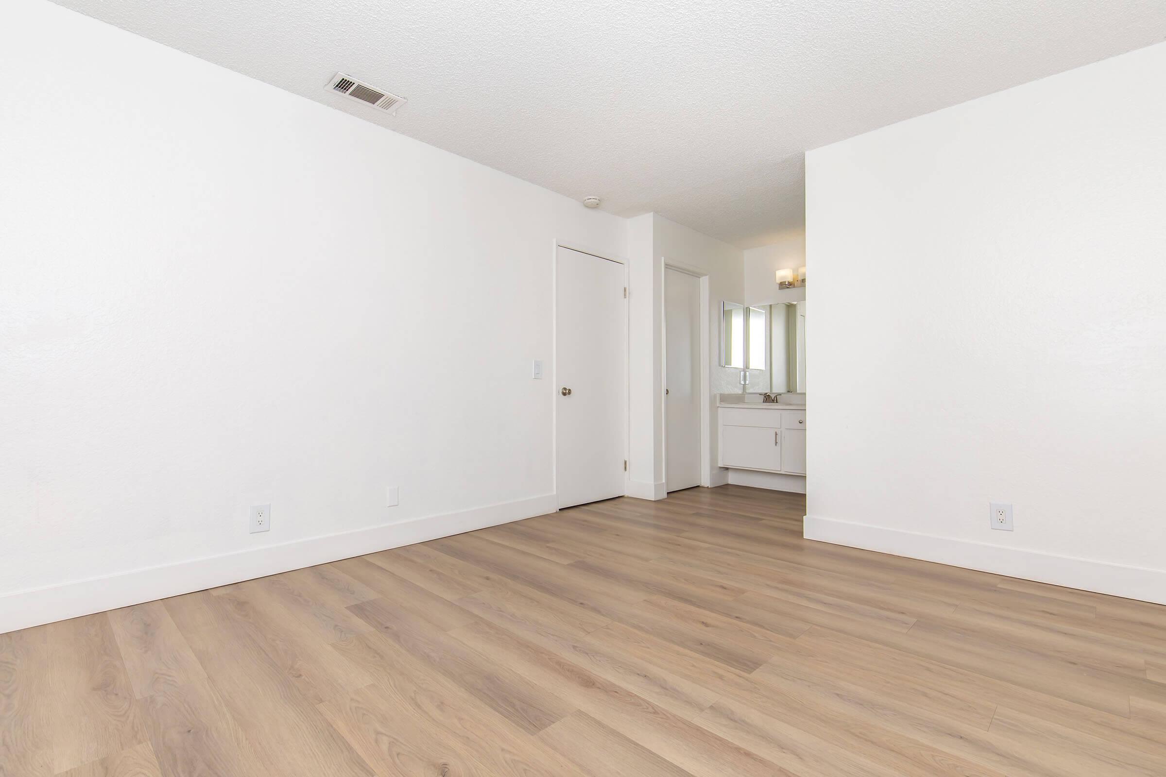 A spacious, empty room with light-colored walls and a light wooden floor. There's a door on the left leading to another space, and a bathroom area is visible in the background with a mirror and vanity, illuminated by a soft light fixture. The overall atmosphere is bright and minimalist.