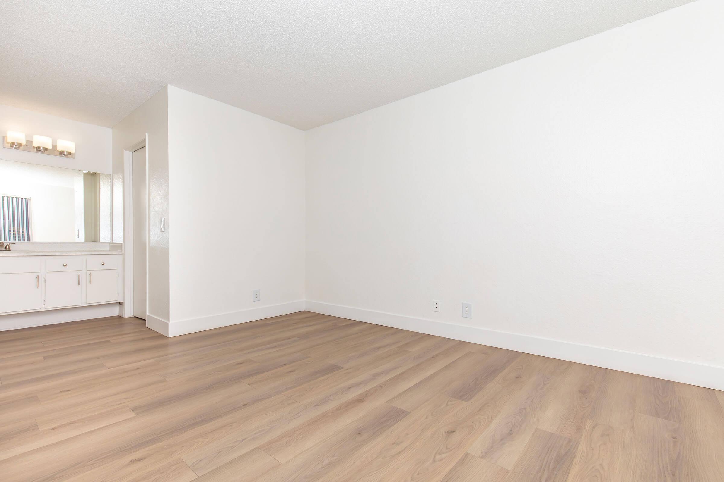 A spacious, empty room with light-colored walls and wooden flooring. The left side features a bathroom area with a mirror and countertop, while the right side is open and minimalist, creating a bright and airy atmosphere.
