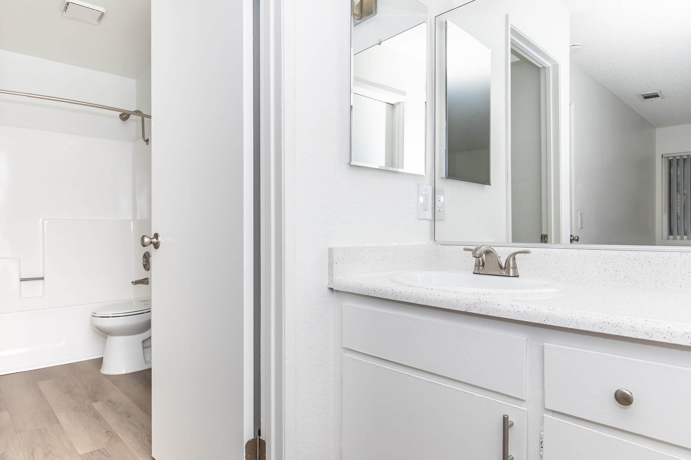 A bright and clean bathroom featuring a white vanity with a sink, two mirrors above the sink, and a light-colored countertop. A door on the left leads to the bathroom area, which includes a shower and toilet. The flooring is light wood, creating a modern and spacious feel.
