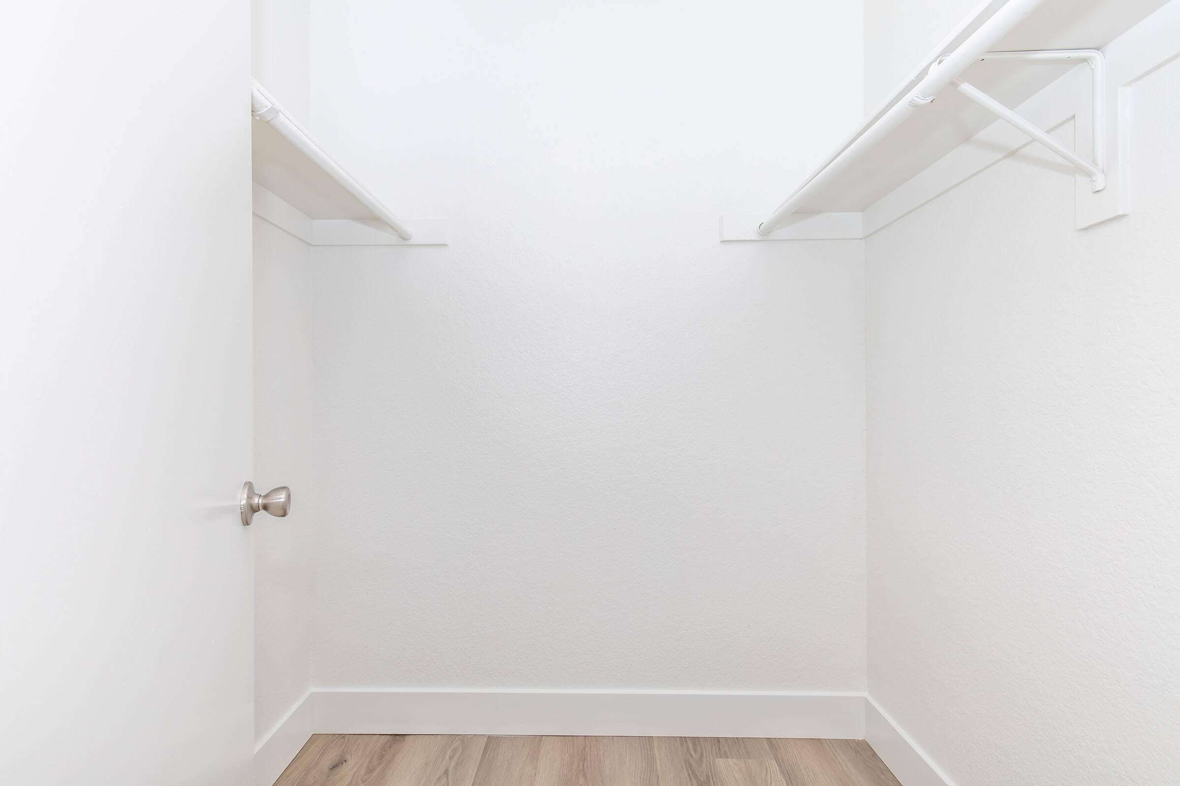 Empty closet with white walls and light wood flooring. Two horizontal shelves are installed on the side walls, and a silver doorknob is visible on the closed door. The space appears clean and minimalistic, creating a sense of openness.