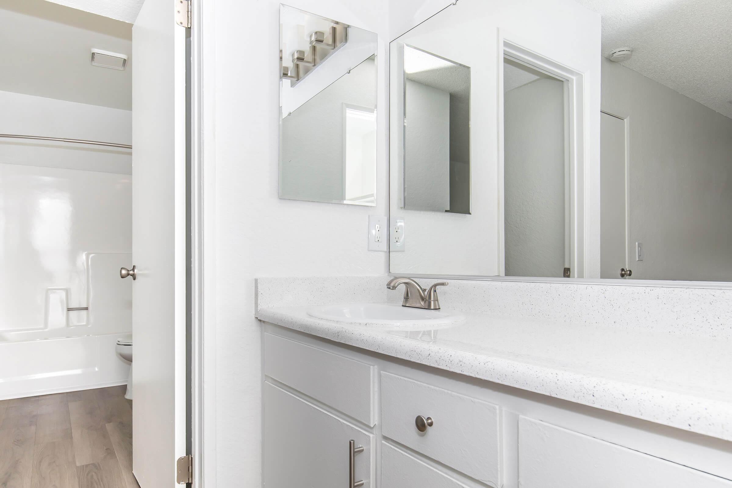 A modern bathroom featuring a white countertop with a sink and faucet, two mirrors above, light-colored walls, and an adjacent bathroom area with a bathtub and shower. The floor is made of light wood laminate, and the room is well-lit with natural light coming from a nearby window.