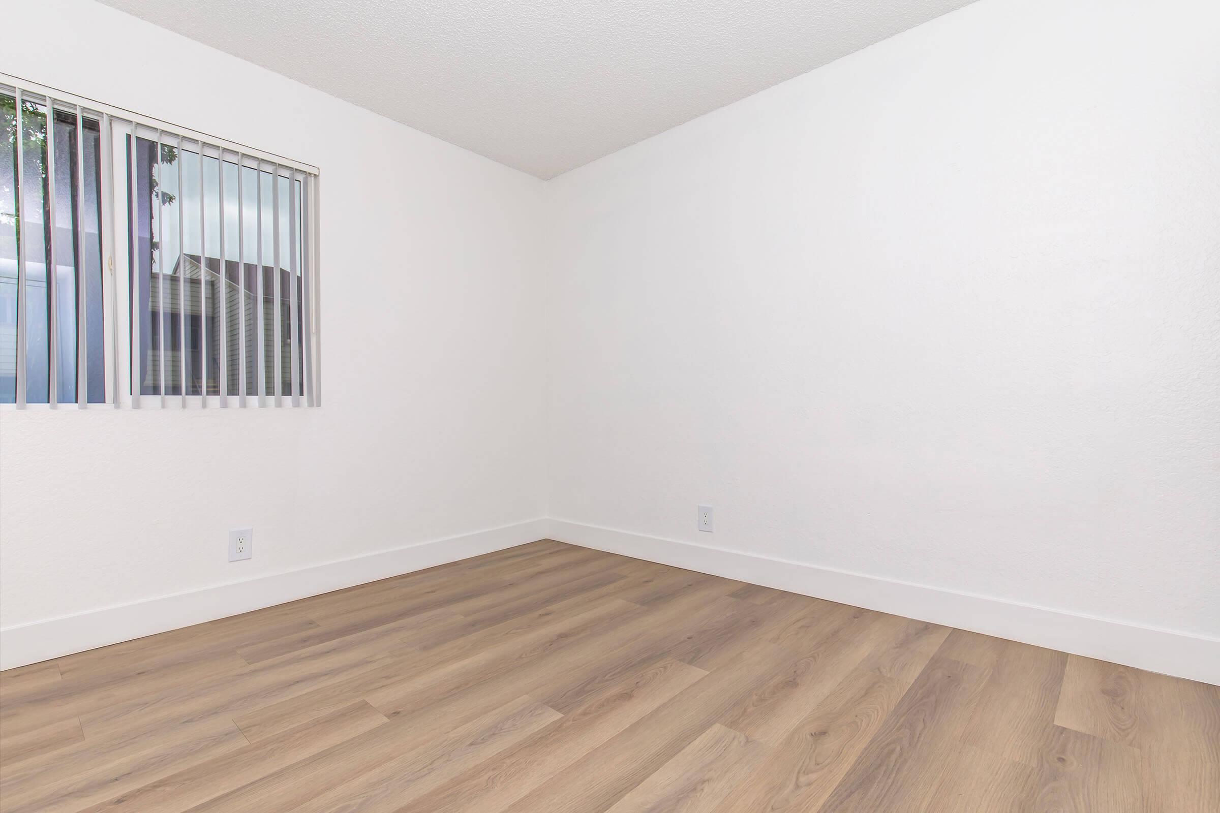 Empty room with light-colored walls, a large window featuring vertical blinds, and wooden flooring. The space is bright and minimalistic, with no furniture present, creating an open and airy atmosphere.