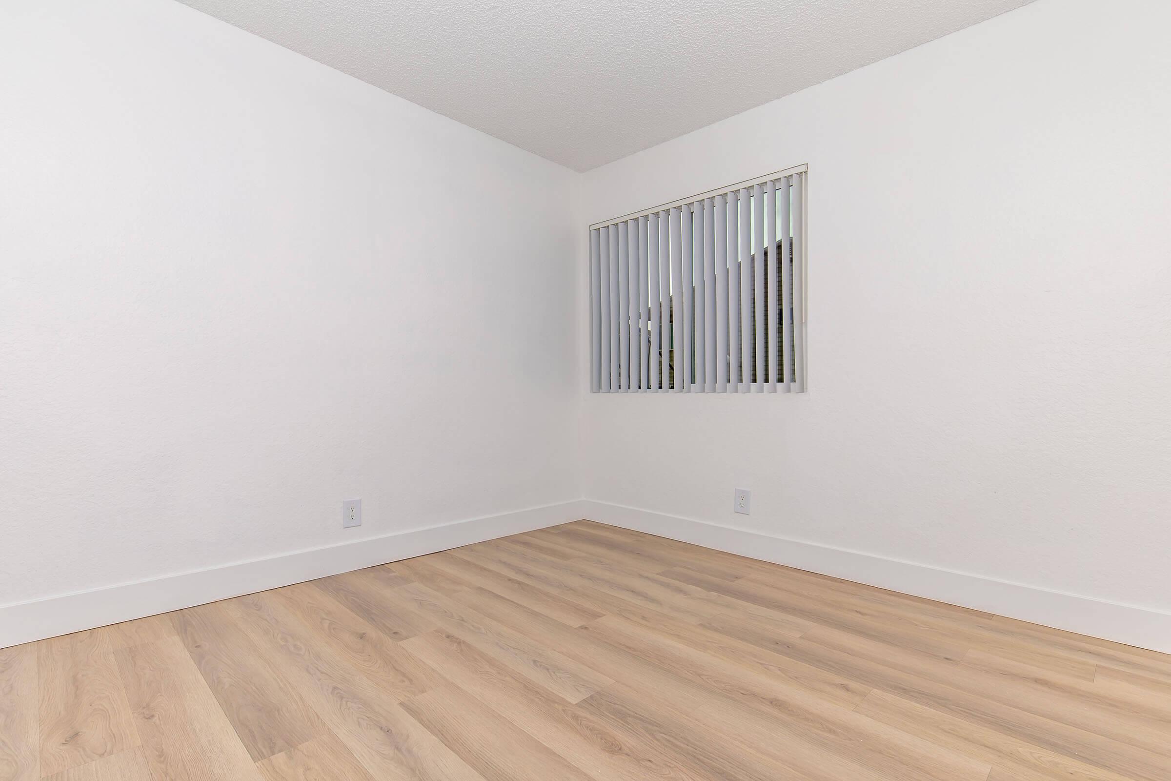 A bright, empty room with light-colored walls and wooden flooring. It features a window with vertical blinds allowing natural light to enter, creating a spacious and minimalist atmosphere suitable for various uses or furnishings.