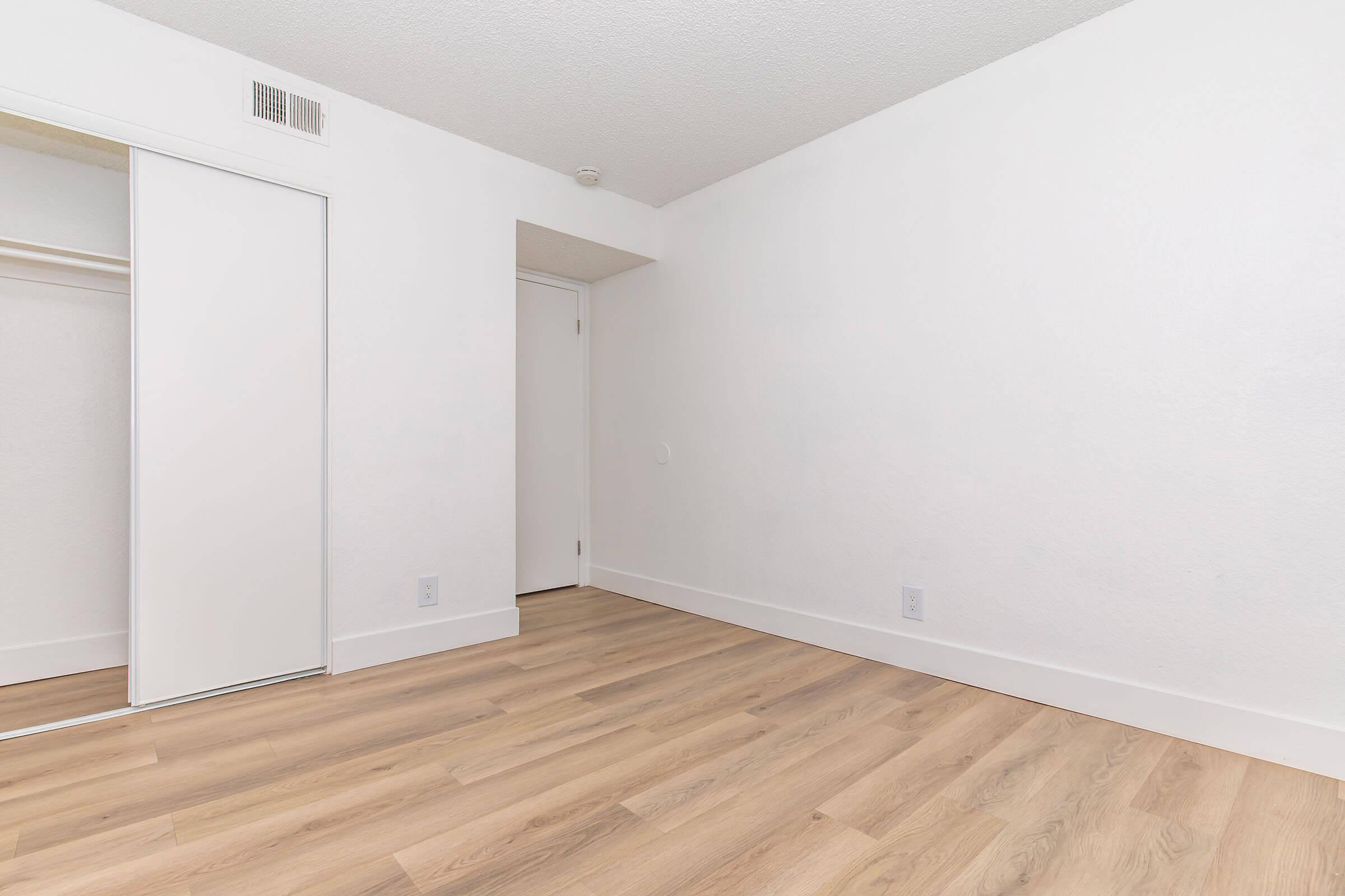 Empty room with light-colored walls and wooden flooring. There's a closet with sliding doors on the left side. A plain white door is visible on the right side, leading to another space. Natural light illuminates the room, giving it a bright and spacious feel.