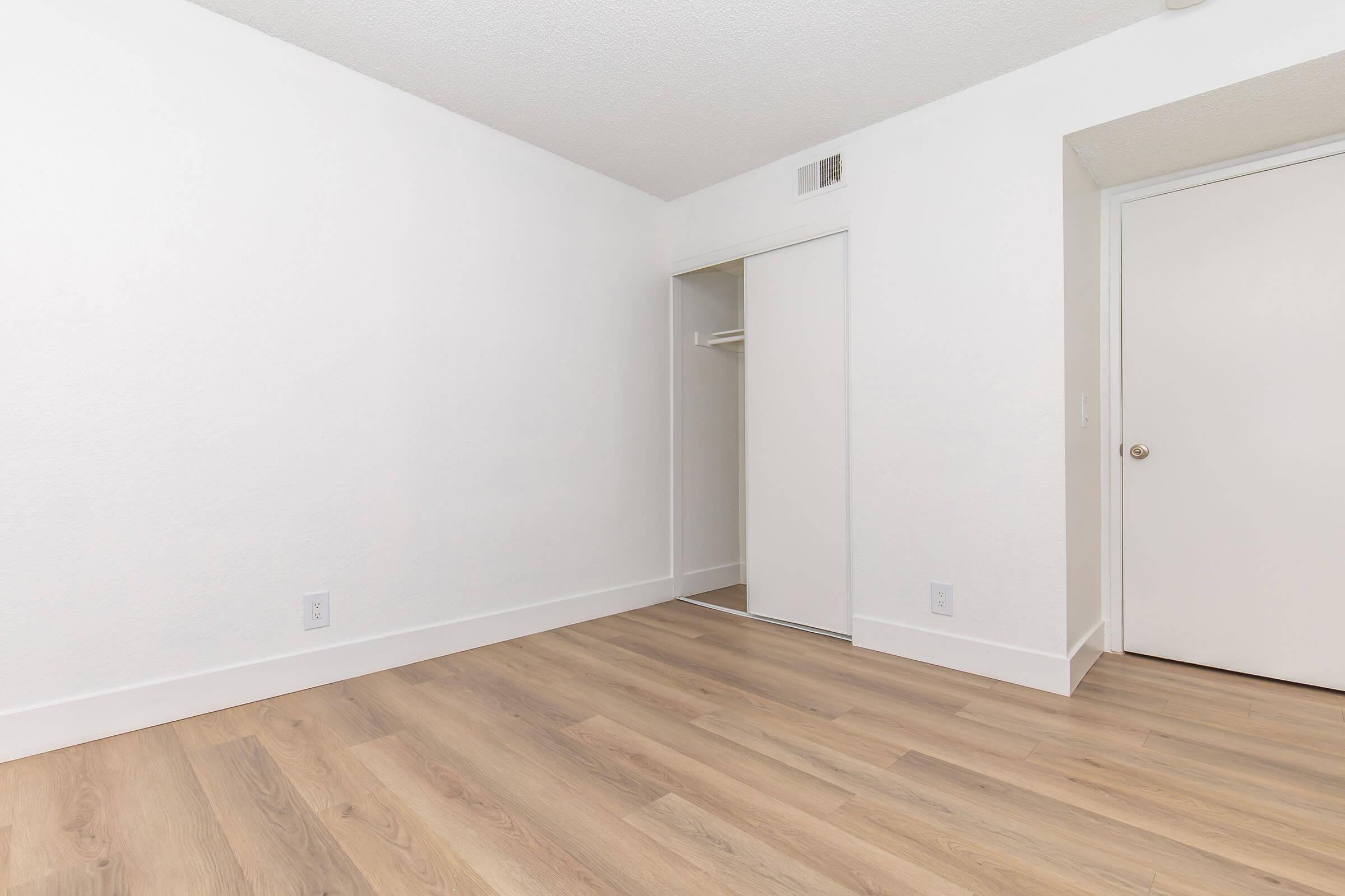 A minimalistic interior of a room featuring white walls and light wood flooring. There is a closet with sliding doors on the left side and a closed door on the right side. The overall ambiance is bright and clean, suggesting a modern and spacious feel.
