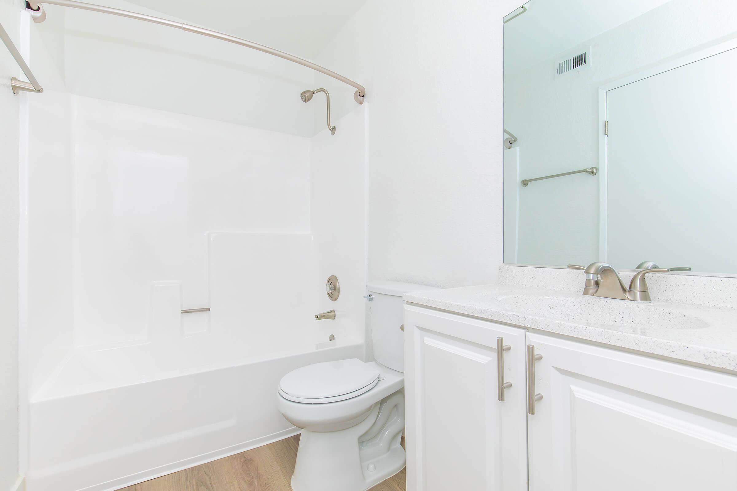 A clean and modern bathroom featuring a white bathtub with a curved shower rod, a white toilet, and a double vanity with a light-colored countertop. The walls are painted white, and a large mirror reflects the space, enhancing the bright and airy feel of the room.