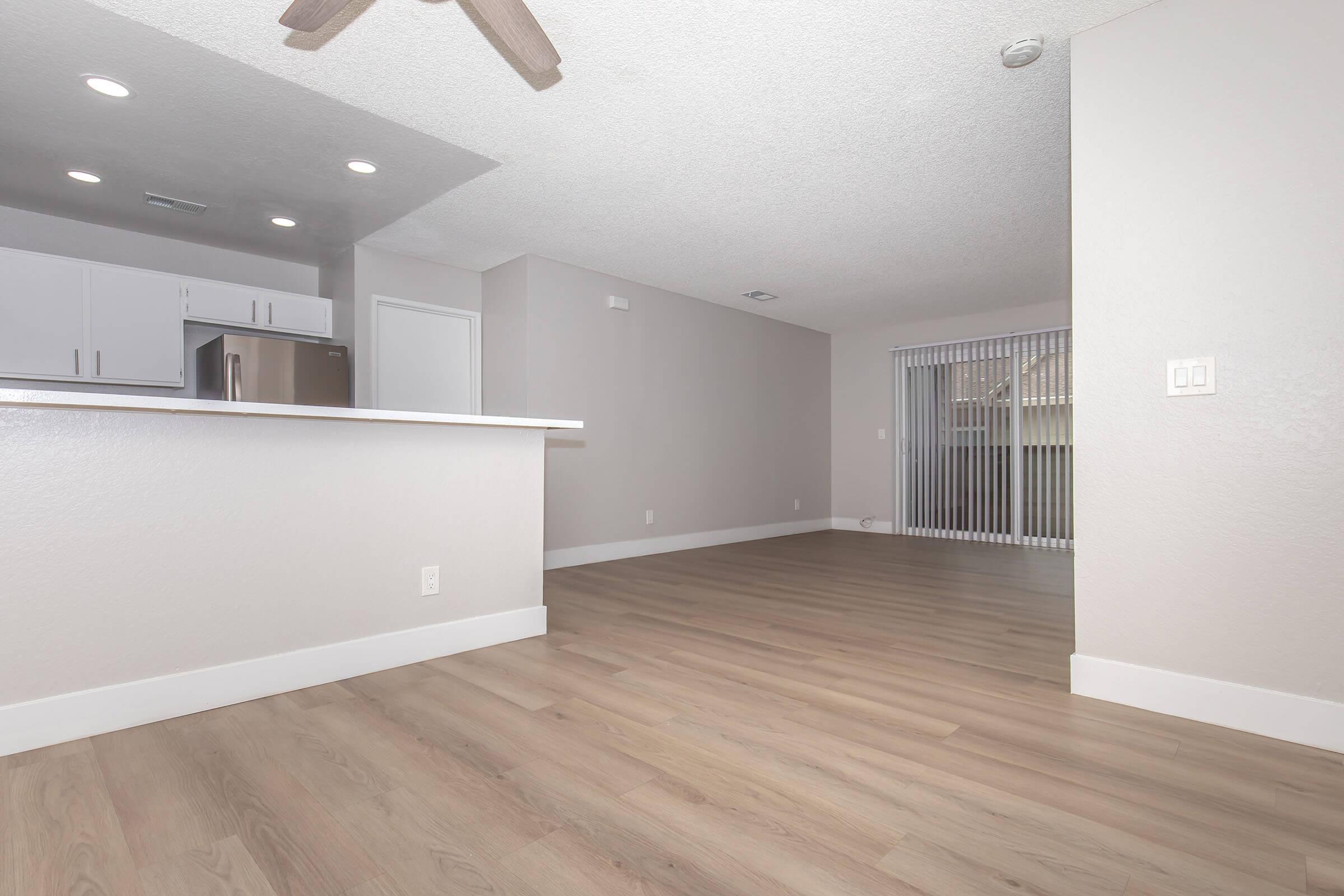 A spacious, light-filled living area with modern finishes. The open layout features a wooden floor, a kitchen island with countertops, and a view of a sliding glass door leading to an outdoor space. The walls are painted in soft gray tones, creating a welcoming atmosphere.
