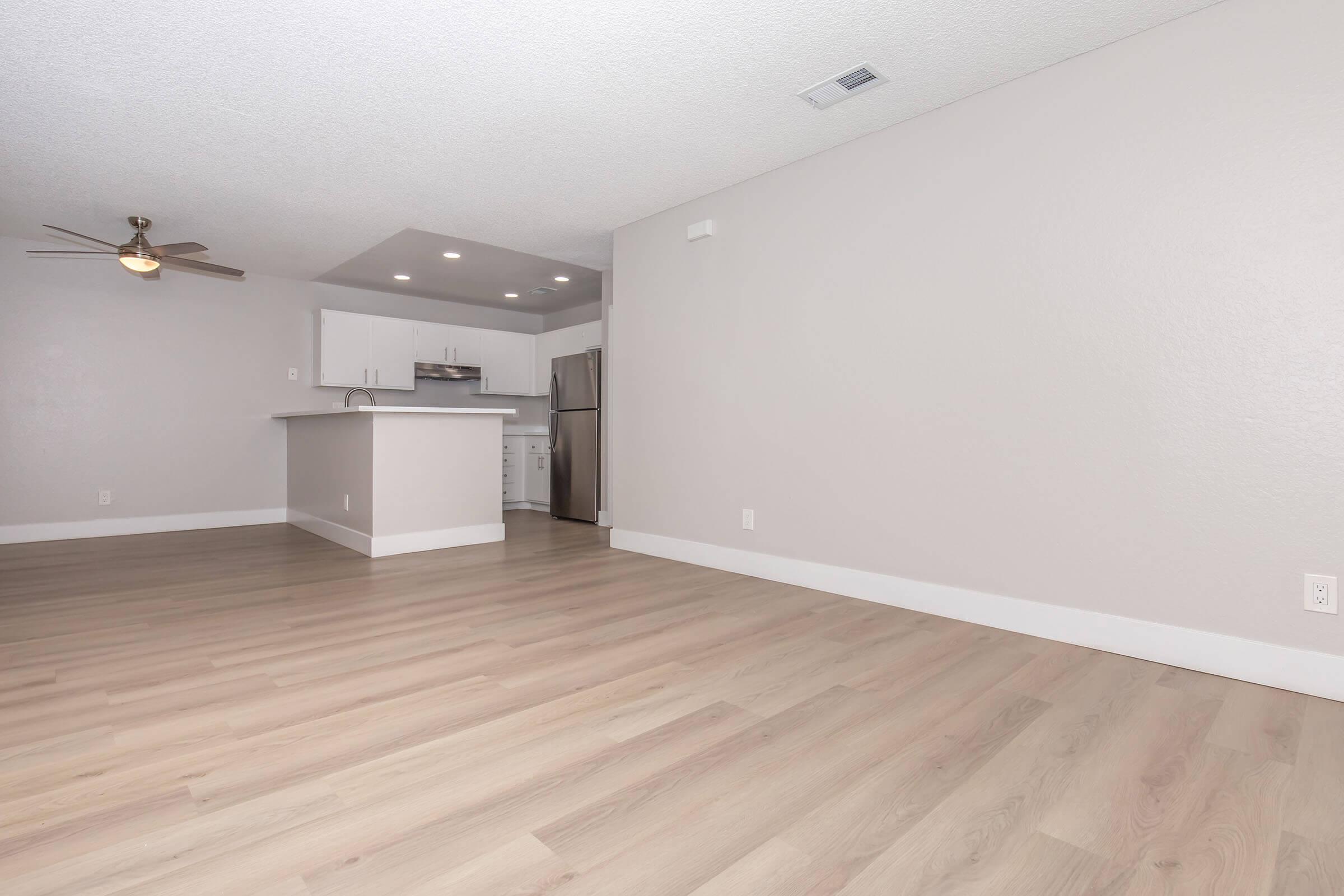 A modern, open-concept living area featuring light-colored walls, a ceiling fan, and a spacious wood laminate floor. The kitchen is visible in the background, equipped with stainless steel appliances. Natural light brightens the space, creating a fresh and inviting atmosphere.