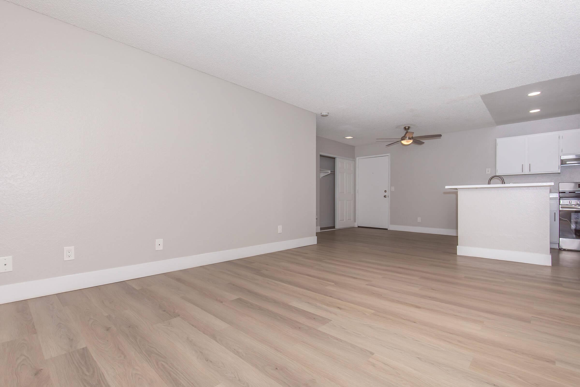 Interior view of a modern, unfurnished living space featuring light-colored walls and hardwood floors. There is a ceiling fan, an open kitchen area in the background with white cabinetry, and a door leading to another room. The space is well-lit and appears spacious.
