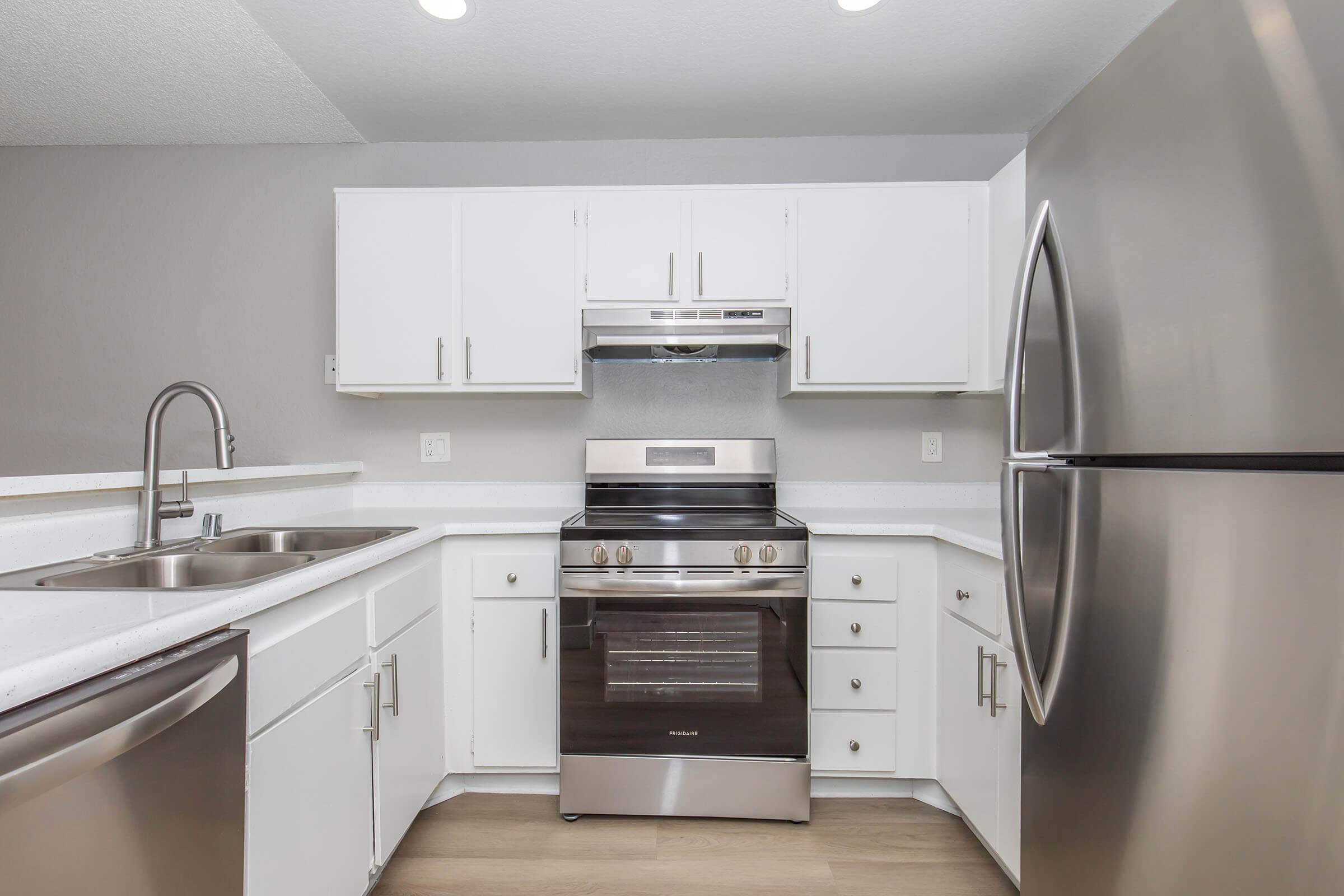 Modern kitchen featuring white cabinetry, stainless steel appliances including a stove and refrigerator, and a double sink. The countertops are light-colored, and the space is well-lit with overhead lighting. The flooring is a light wood finish, creating a clean and contemporary look.