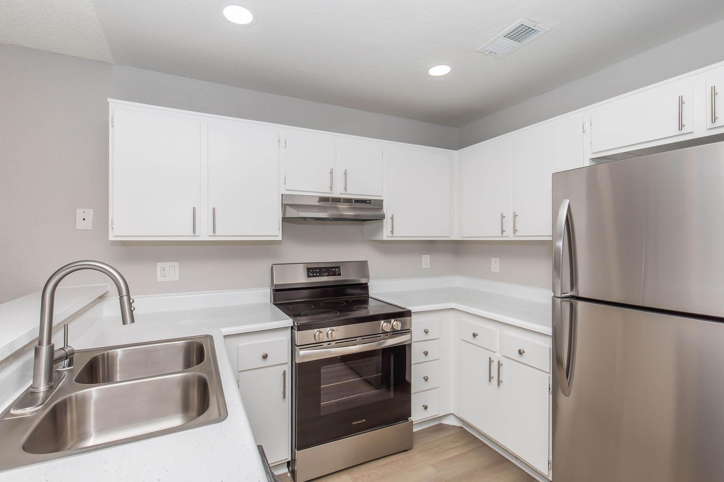 A modern kitchen featuring white cabinets, stainless steel appliances including a stove and refrigerator, and a double sink. The countertops are light-colored with a smooth finish, and the walls are painted in a neutral tone, creating a clean and bright ambiance.