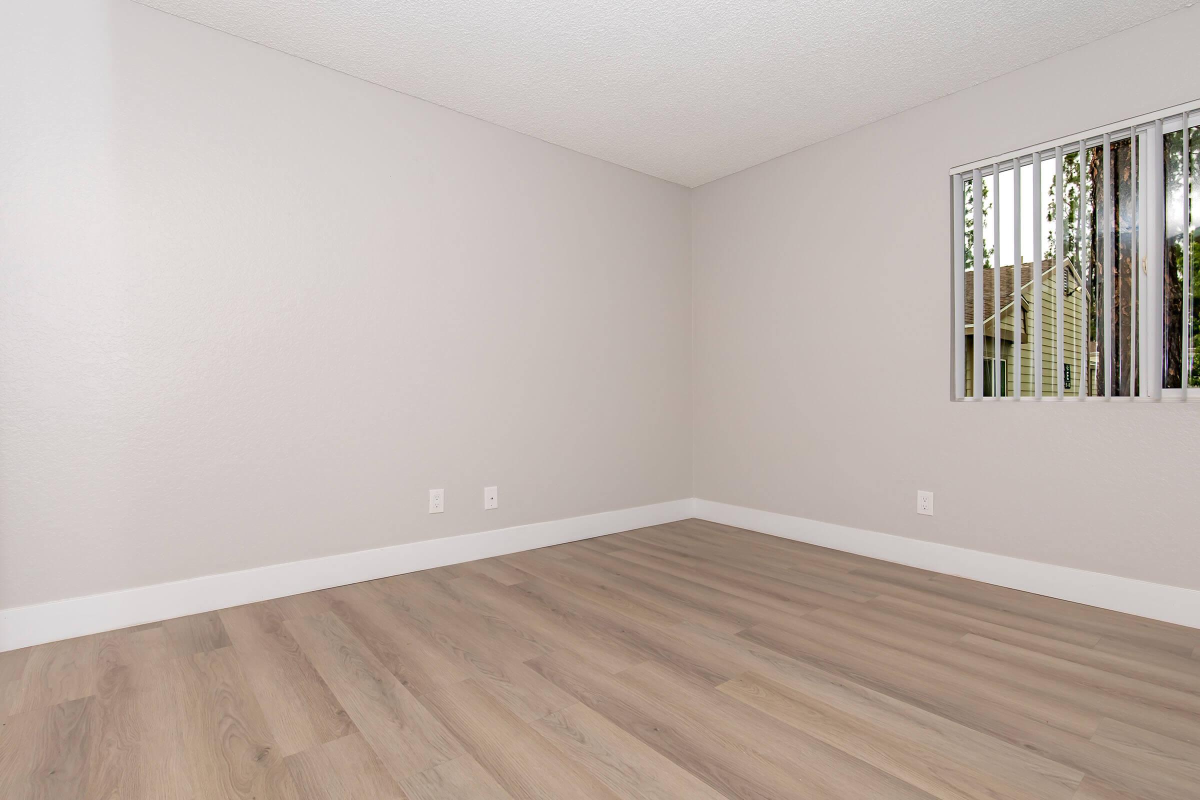 Empty room with light gray walls and laminate flooring. A narrow window with horizontal blinds allows natural light to enter, revealing greenery outside. The space appears clean and minimalistic, suitable for various uses or décor.