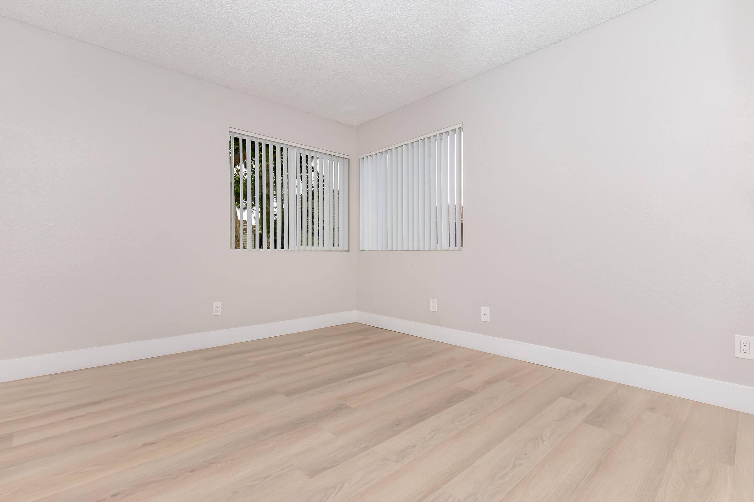 Empty room with light grey walls and pale wooden floors. Two windows with vertical blinds allow natural light to enter. The space is untidy and minimalistic, featuring no furniture or decorations.