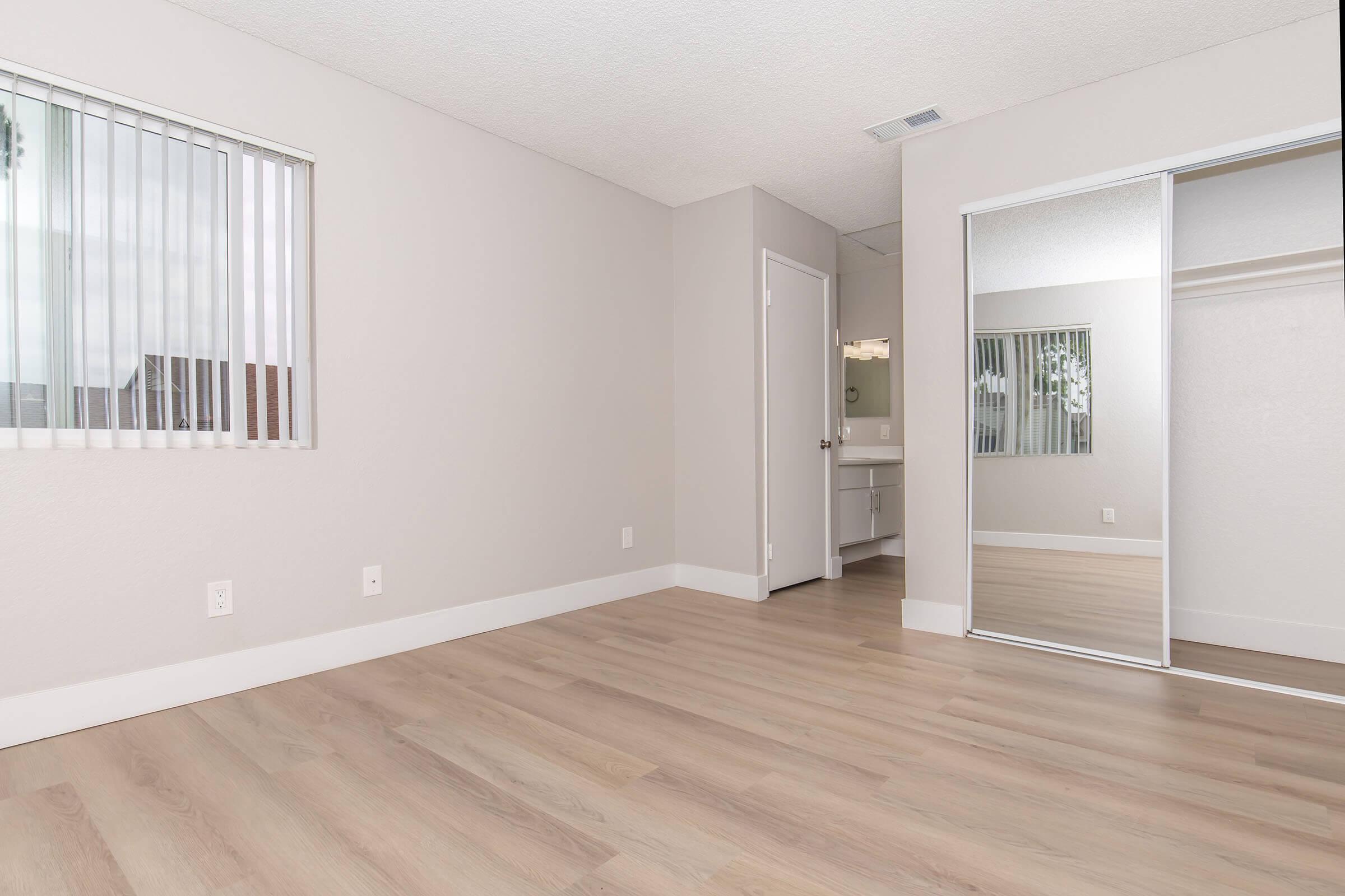A spacious, minimally furnished room with light-colored walls and laminate flooring. Large windows with vertical blinds allow natural light in. A mirrored closet door reflects part of the room. A doorway leads to a bathroom area, enhancing the open feel of the space.