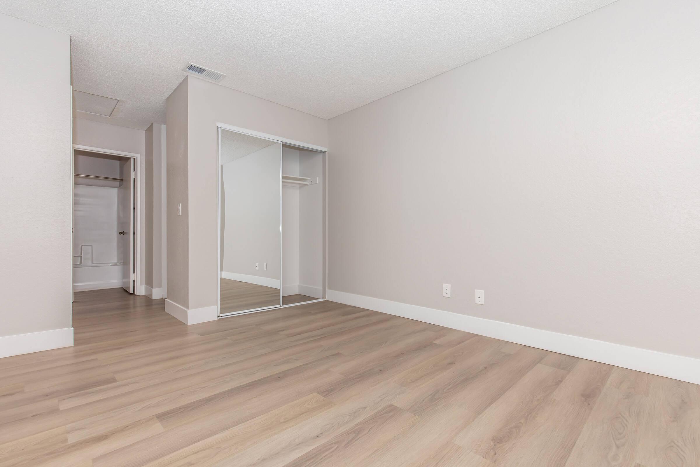 Empty room with light gray walls and laminate flooring. A large mirror-door closet is on one side, and a doorway leads to a bathroom with a shower visible. Natural light fills the space, creating a bright and open atmosphere.