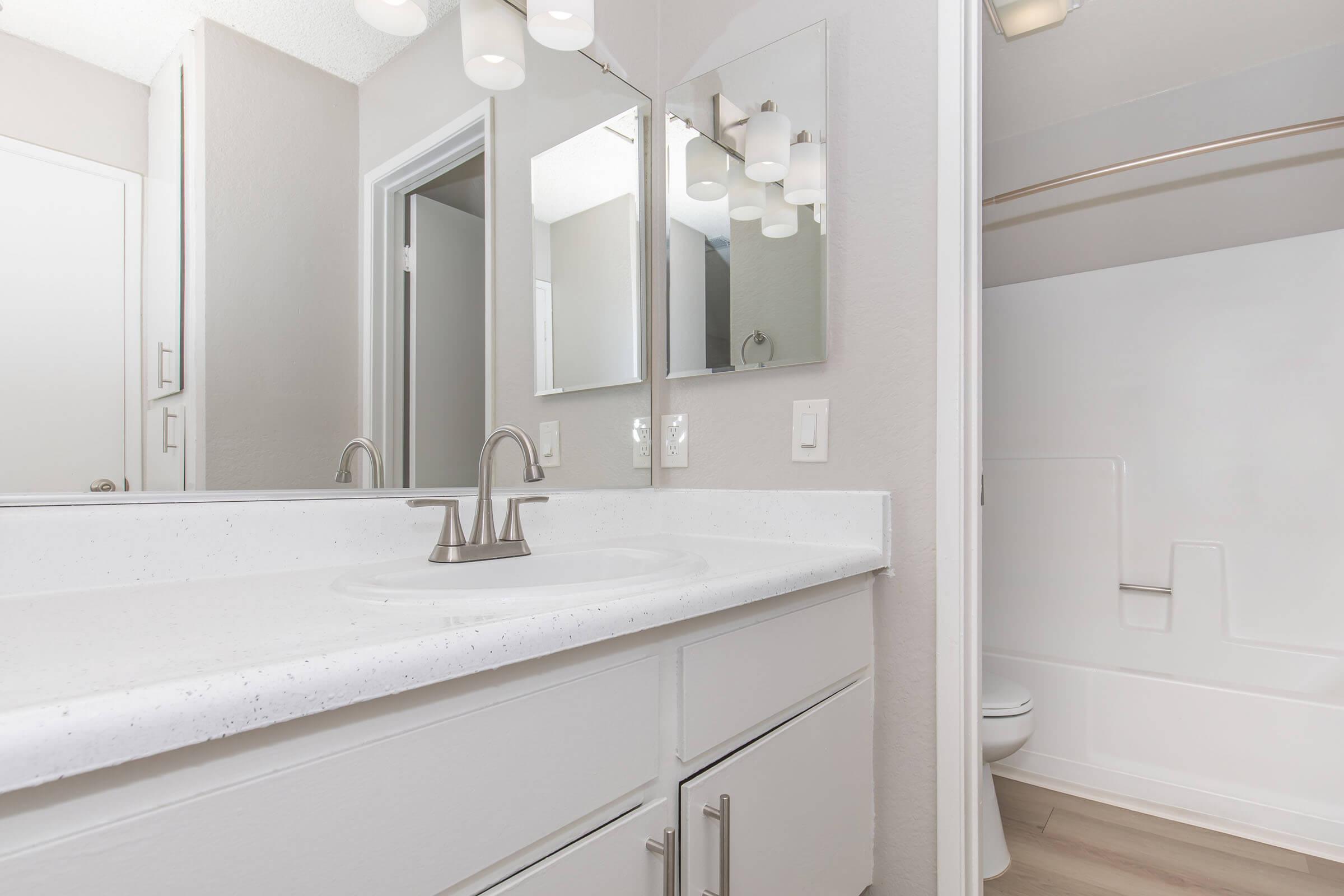A modern bathroom featuring a double sink vanity with a white countertop, two mirrors, and stylish light fixtures. The walls are painted a light gray, and there is a bathtub with a shower curtain visible in the background. The flooring is light-colored wood laminate.