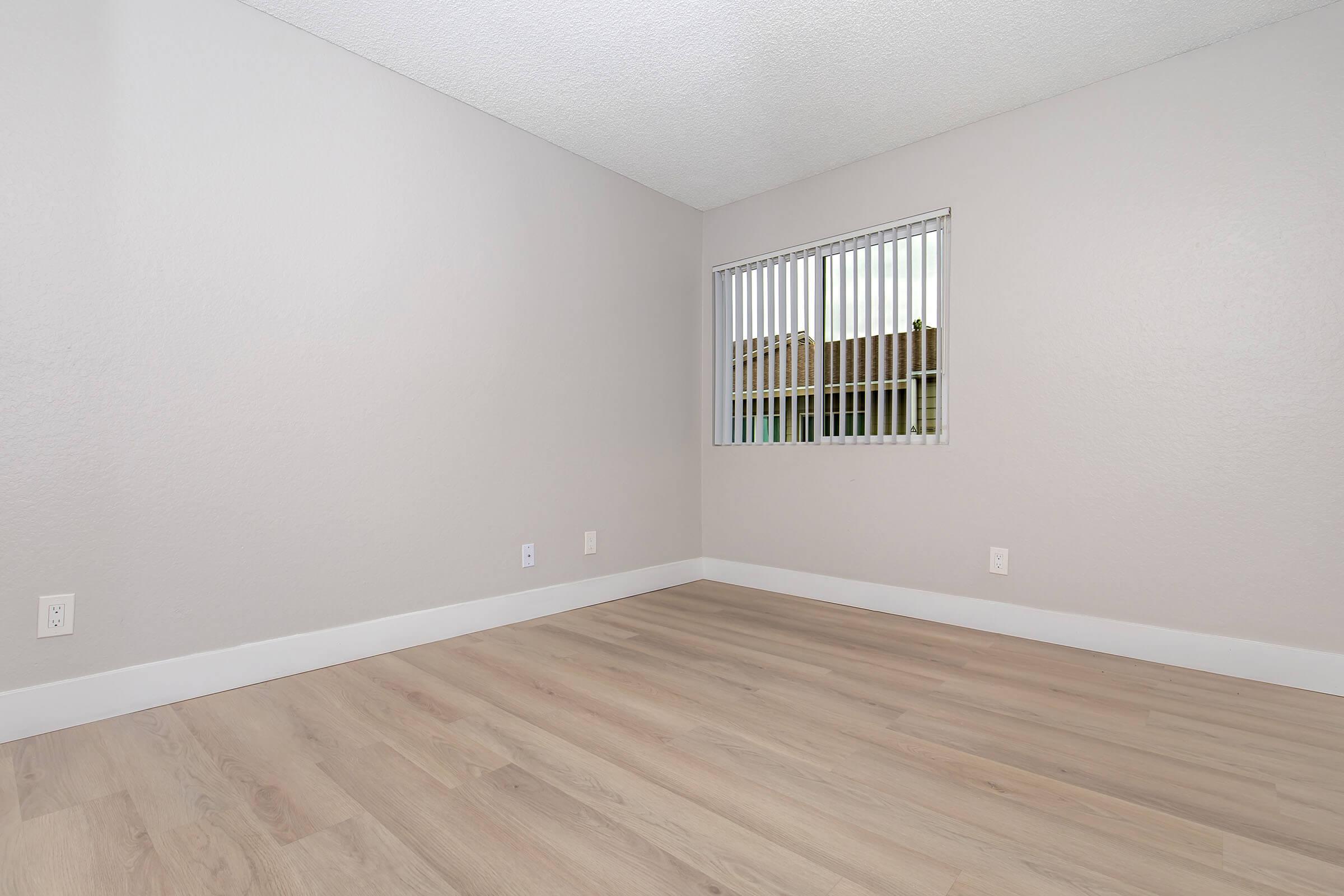 A minimalist interior of a room featuring light-colored walls and a window with vertical blinds. The floor is made of light wooden planks, creating a spacious and airy feel. Natural light is entering through the window, illuminating the uncluttered space.