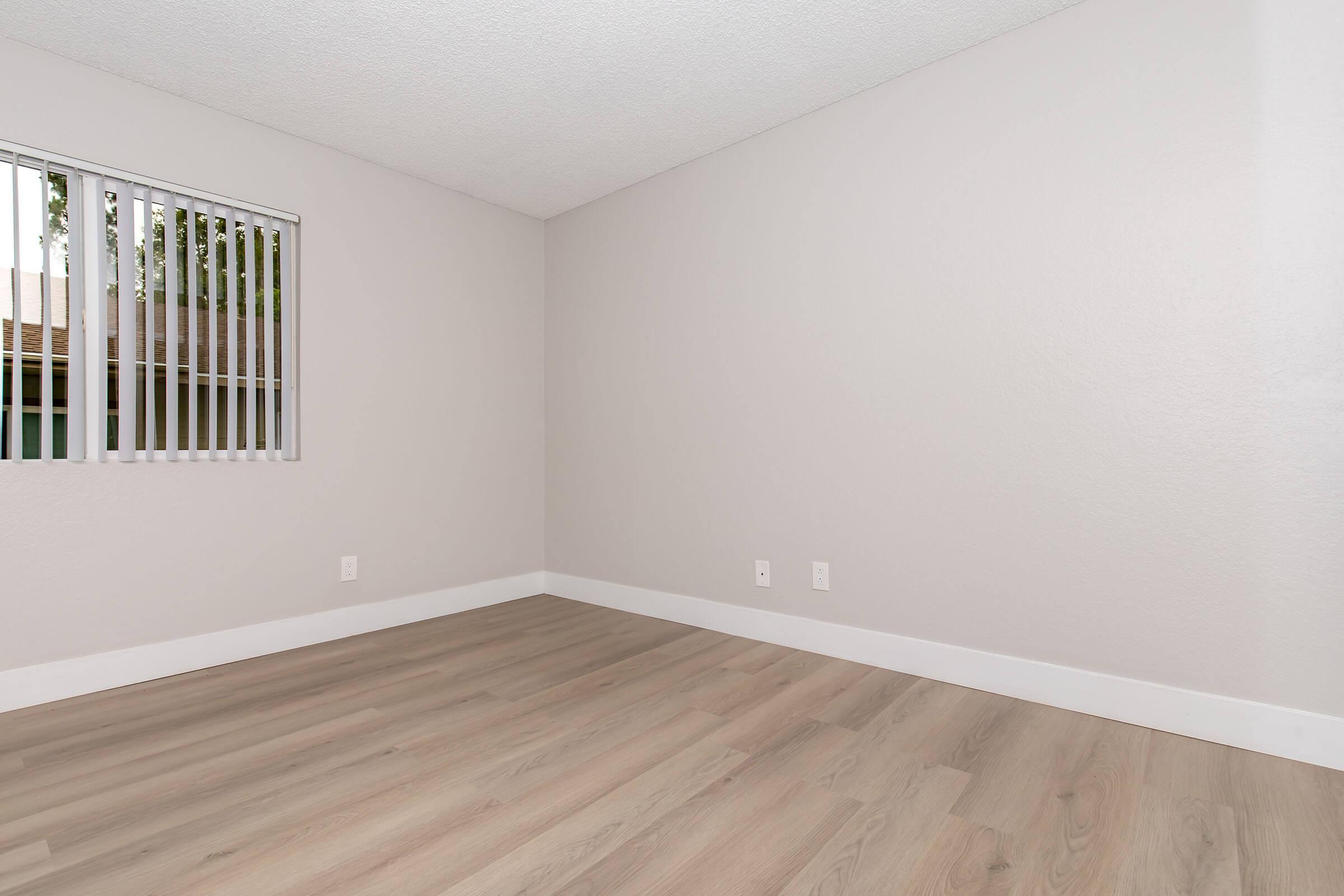 A clean, empty room with light gray walls and light wooden flooring. There are vertical blinds on a window letting in natural light, and the room appears spacious and minimalistic. No furniture or decorations are present.