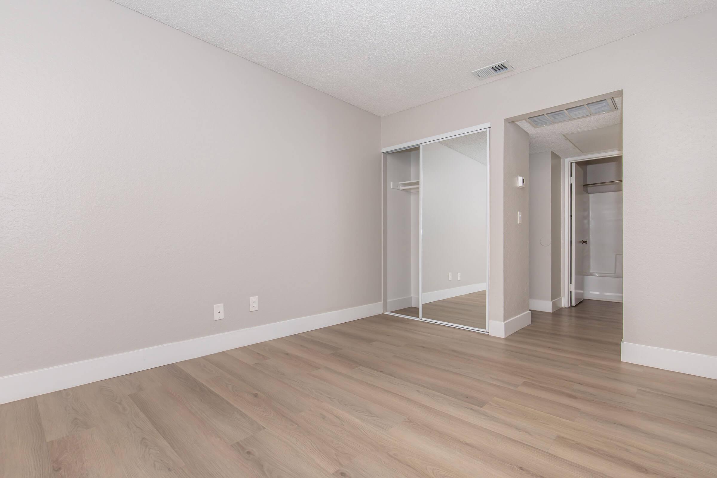 A spacious, empty room with light gray walls and wood-look laminate flooring. There is a mirrored closet on one side and an open area leading to a bathroom in the background. The lighting is bright and the overall atmosphere is clean and modern.