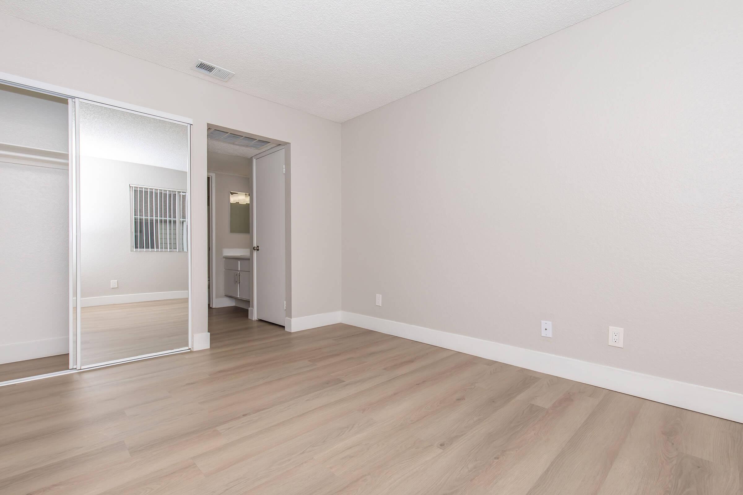 A spacious, empty room with light-colored walls and wooden flooring. A mirrored sliding closet door is visible on the left, and a doorway leads to an adjacent room. Natural light enters from a window, contributing to a bright and airy atmosphere.