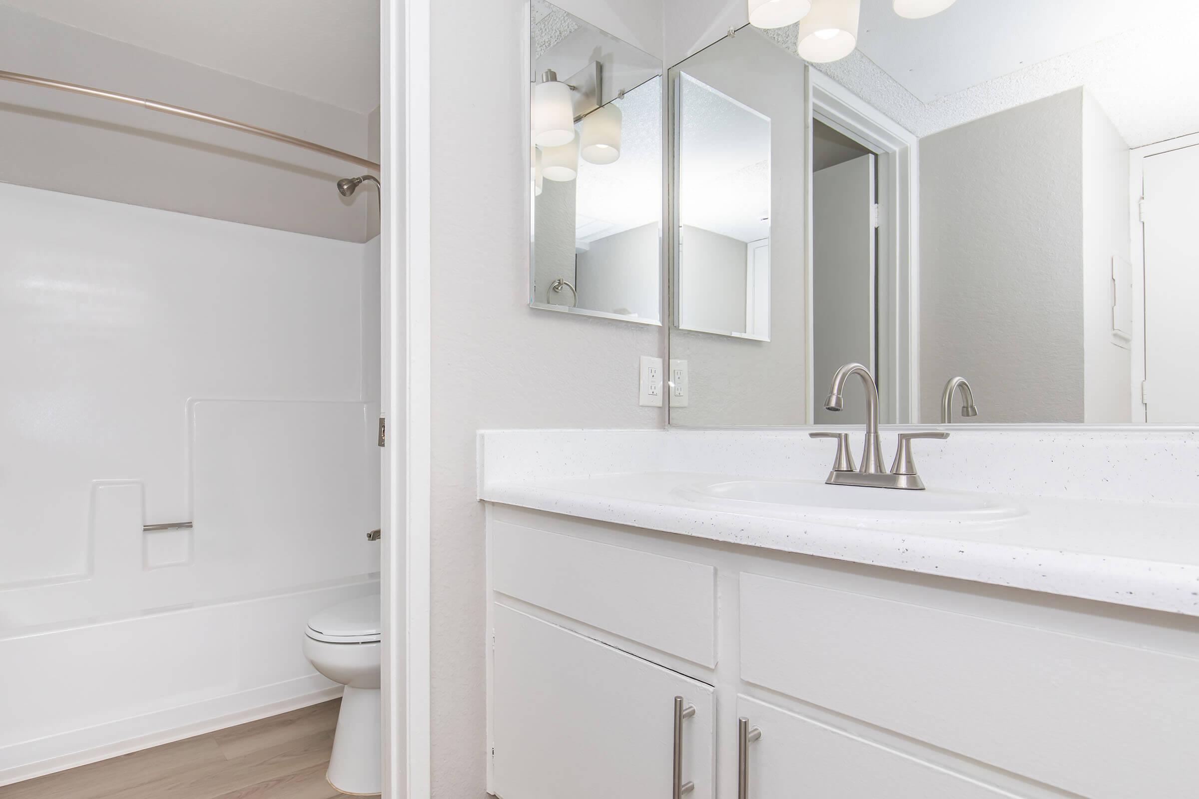 A clean and modern bathroom featuring a white bathtub and shower combo, a sleek vanity with a single sink, and a large mirror. The walls are painted in a light color, enhancing the bright and airy feel of the space. The flooring is light wood, complementing the contemporary design.