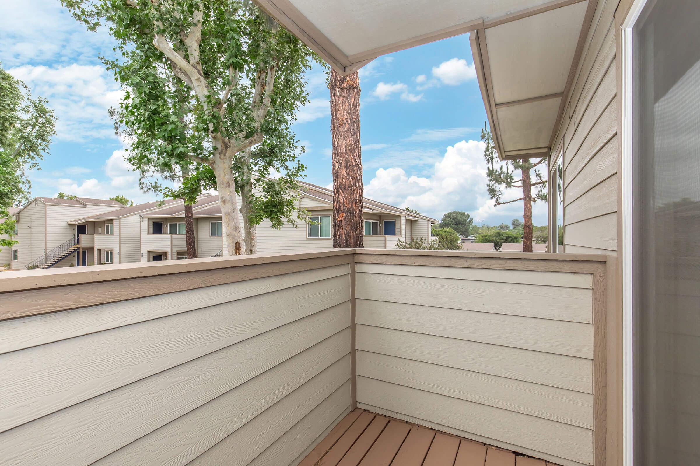 View from a balcony showcasing light-colored wooden railing and flooring. In the background, apartment buildings are visible among green trees under a partly cloudy sky. The scene conveys a serene outdoor space, ideal for relaxation.