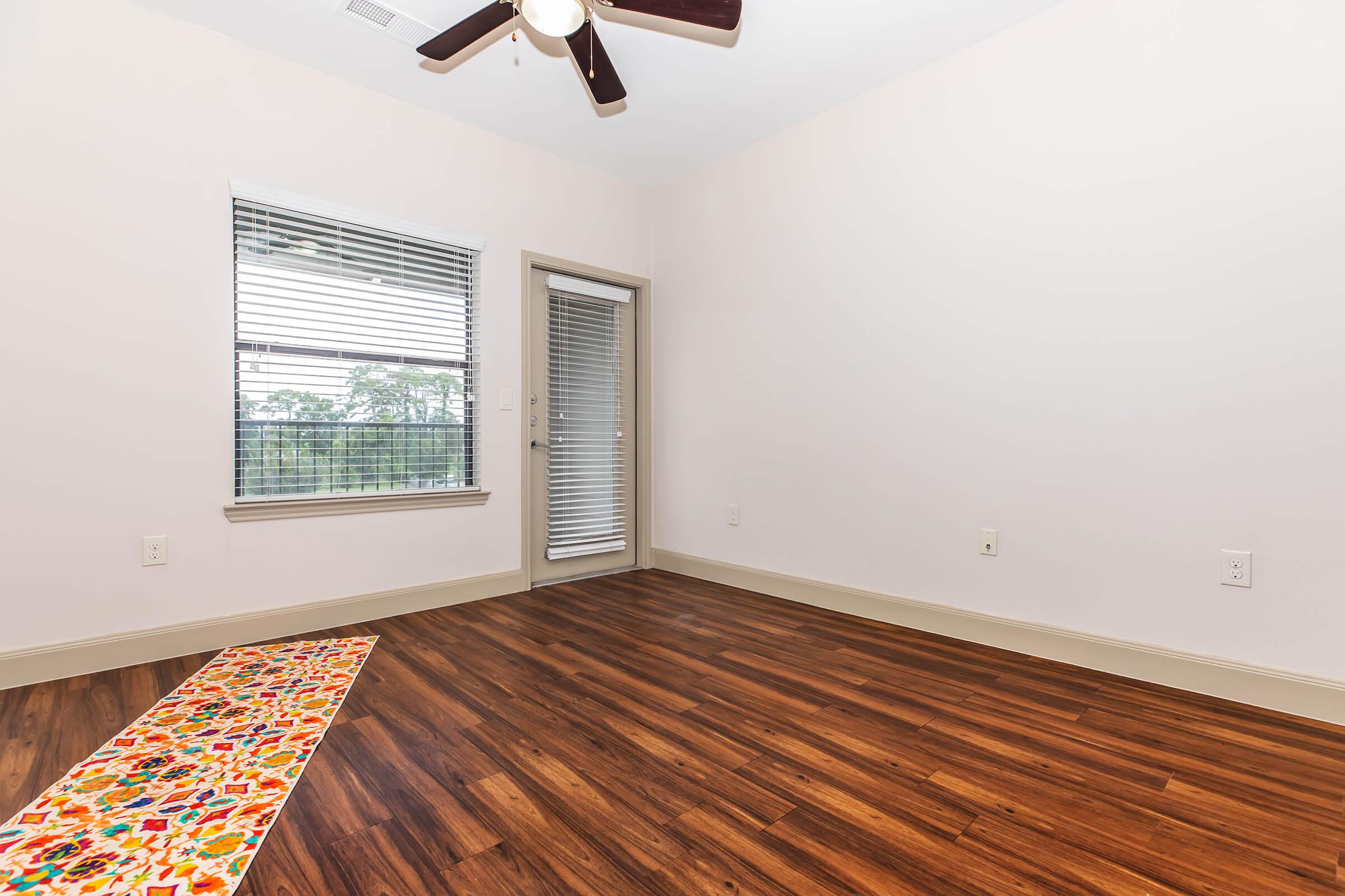 Empty room with light-colored walls, a ceiling fan, and hardwood flooring. A window with blinds allows natural light, and a sliding glass door leads outside. A colorful, patterned runner rug is placed on the floor, adding a touch of color to the otherwise minimalistic space.