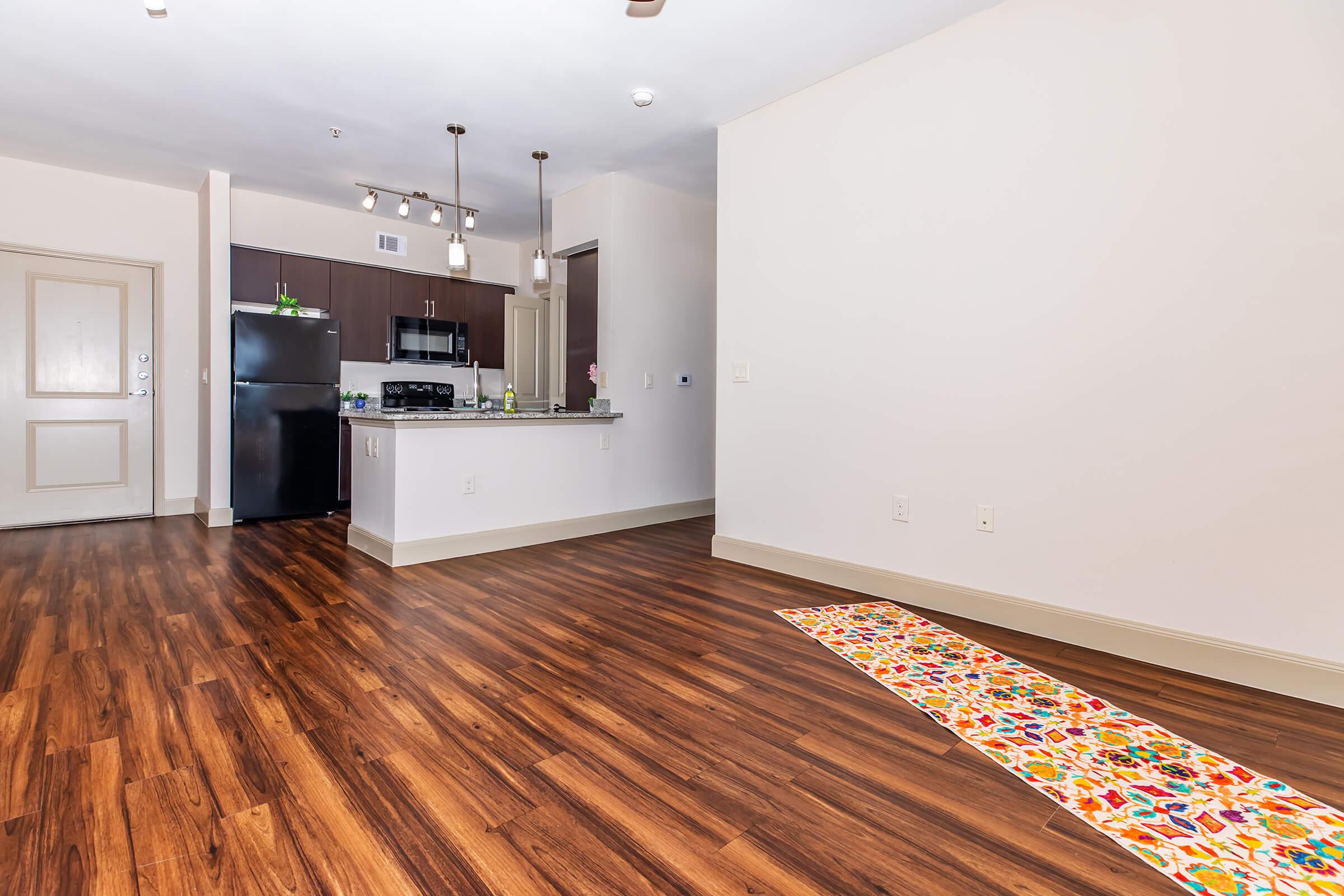 A spacious interior of an apartment featuring hardwood flooring. On the left, a kitchen area with modern black appliances, including a fridge and stove. To the right, a bright, open space leading to a wall with no furniture. A colorful runner rug is placed along the floor, adding a decorative touch.