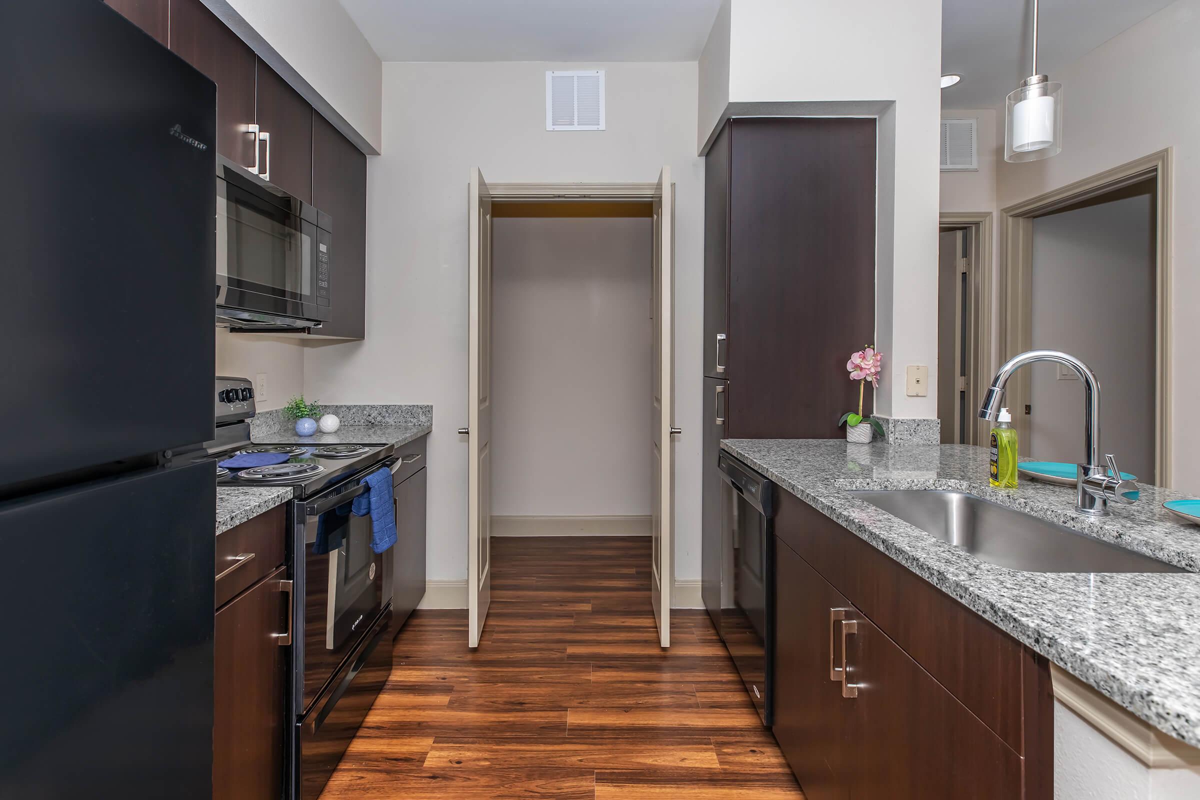 Modern kitchen featuring dark wood cabinetry, granite countertops, and stainless steel appliances. Includes a refrigerator, oven, microwave, and a sink with a faucet. A closet is visible through an open door on the right. The floor is hardwood, and there is a small decorative plant on the countertop.