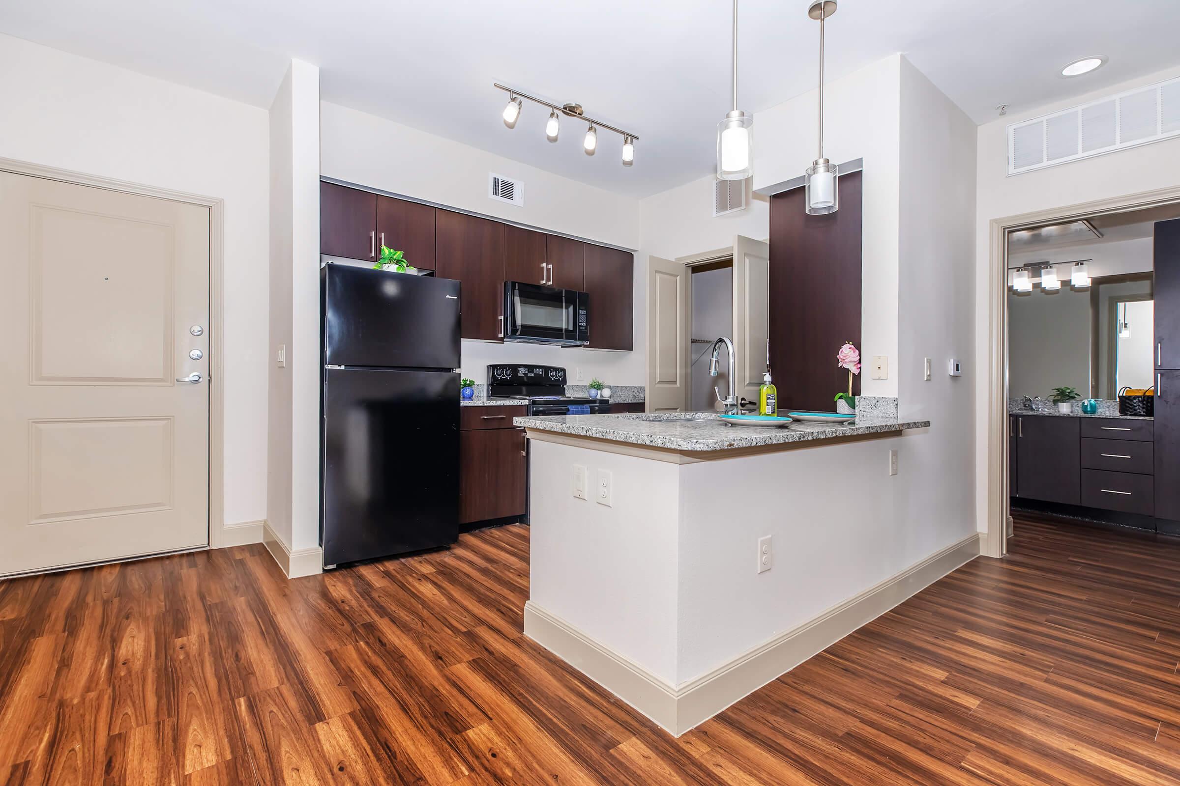 Modern kitchen with dark wood cabinets, granite countertops, and stainless steel appliances. A black refrigerator is visible, along with a microwave and sink. Pendant lights hang above the kitchen island, and the floor features rich brown wood laminate. Light-colored walls create a bright atmosphere.