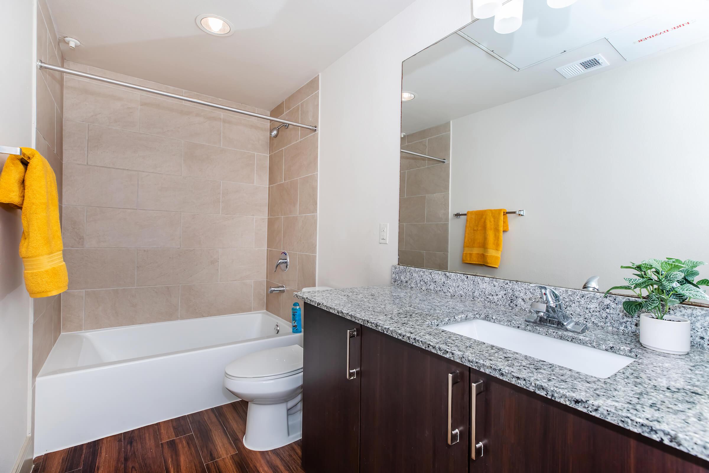 Modern bathroom featuring a bathtub with beige tiles, a sleek granite countertop with a sink, a white toilet, and a large mirror. A bright yellow towel is hanging on a rail, adding a pop of color, and a small potted plant is placed on the countertop for a fresh touch.