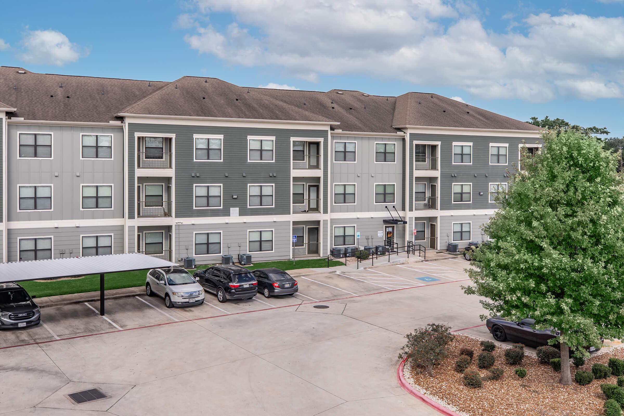 A three-story apartment building with a modern design, featuring gray and white exteriors. Several vehicles are parked in the lot in front, with some covered by carports. Green grass and trees are visible in the surrounding area, and the sky is partly cloudy.