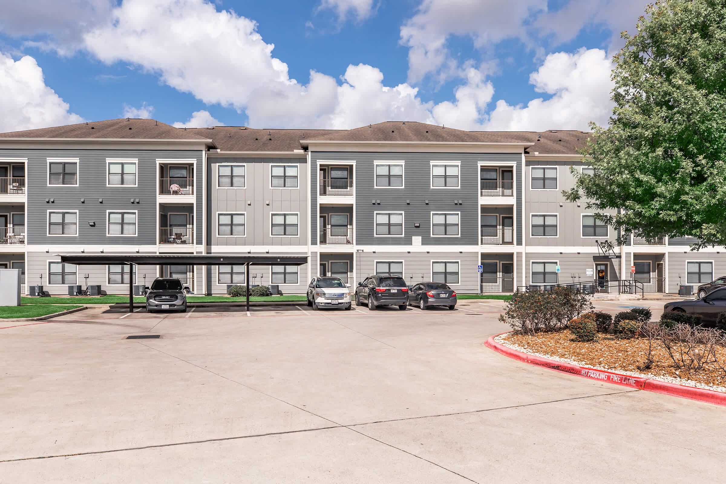 A modern multi-story apartment building with several balconies. In the foreground, there is a parking area with cars under a canopy. The sky is partly cloudy, and there are trees and landscaped shrubs around the building, creating a welcoming residential environment.