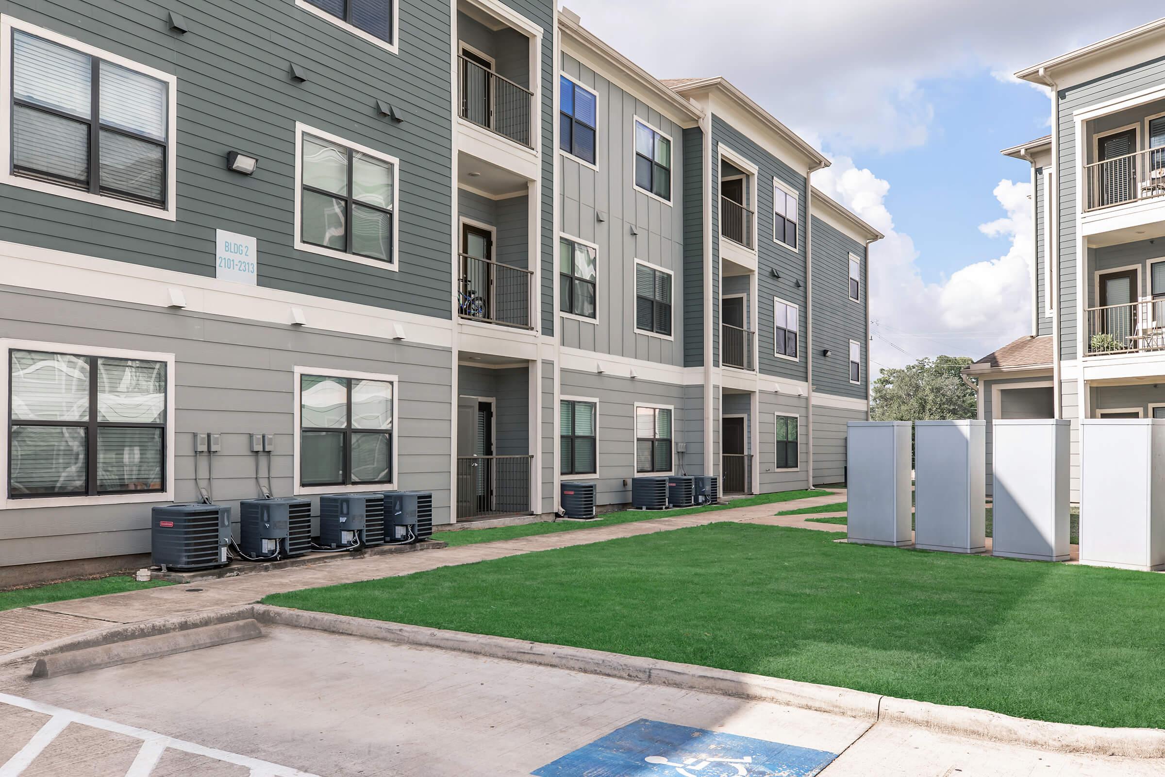 A view of a modern apartment complex featuring three-story buildings with gray exteriors, large windows, and air conditioning units. A well-maintained lawn area with artificial turf is visible in front, along with a parking space marked for accessibility. The sky is partly cloudy.