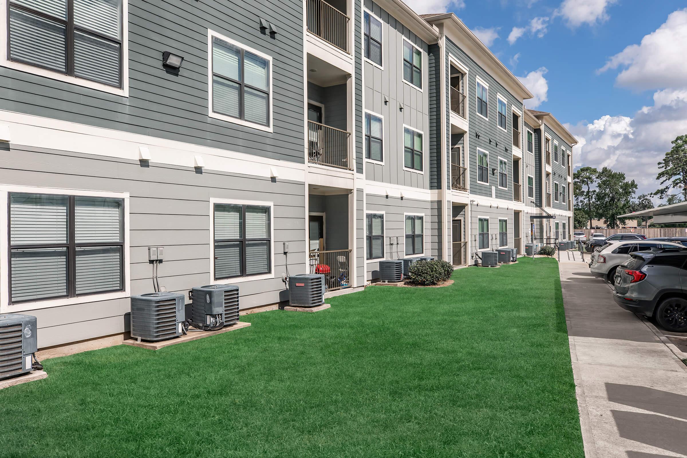 A modern residential apartment building with three stories. The exterior features gray siding and large windows. Green grass is maintained in the foreground, and several air conditioning units are placed near the building. A parking area with cars is visible on the right side. The sky is partly cloudy.