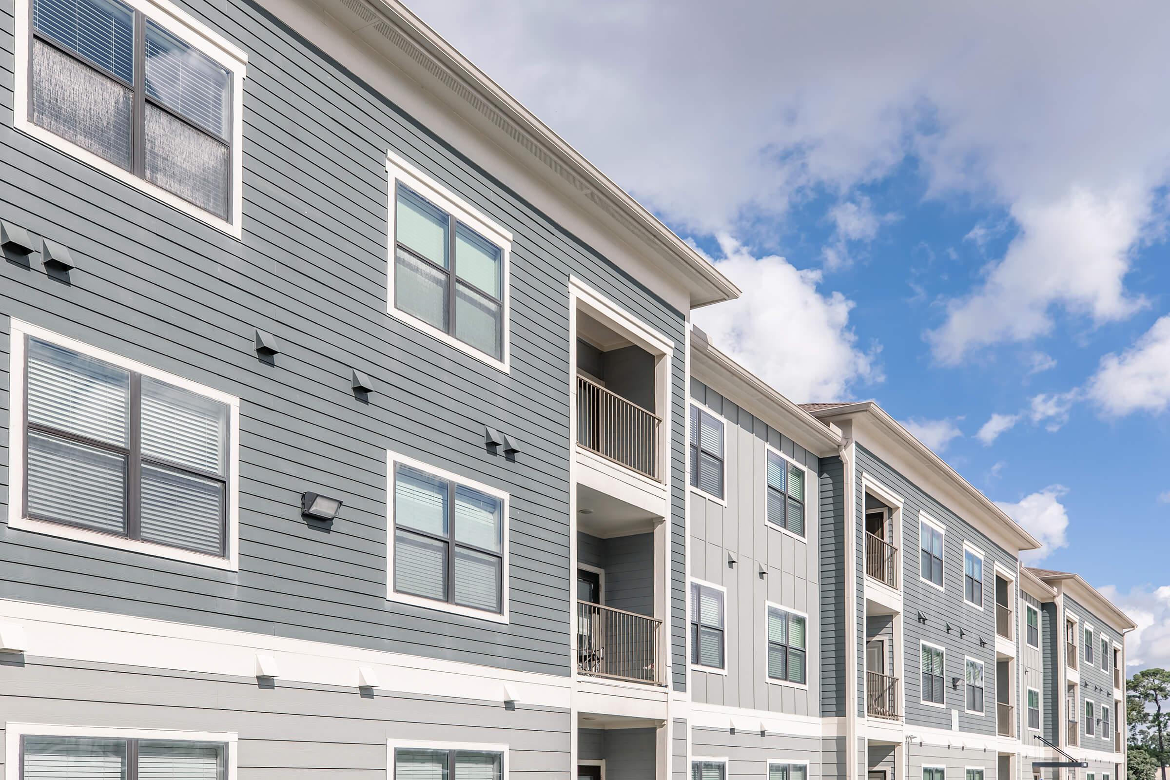 A modern multi-story apartment building with gray siding and white trim. The facade features multiple balconies, large windows, and a clear blue sky with some clouds in the background. The design emphasizes contemporary architecture and residential living.
