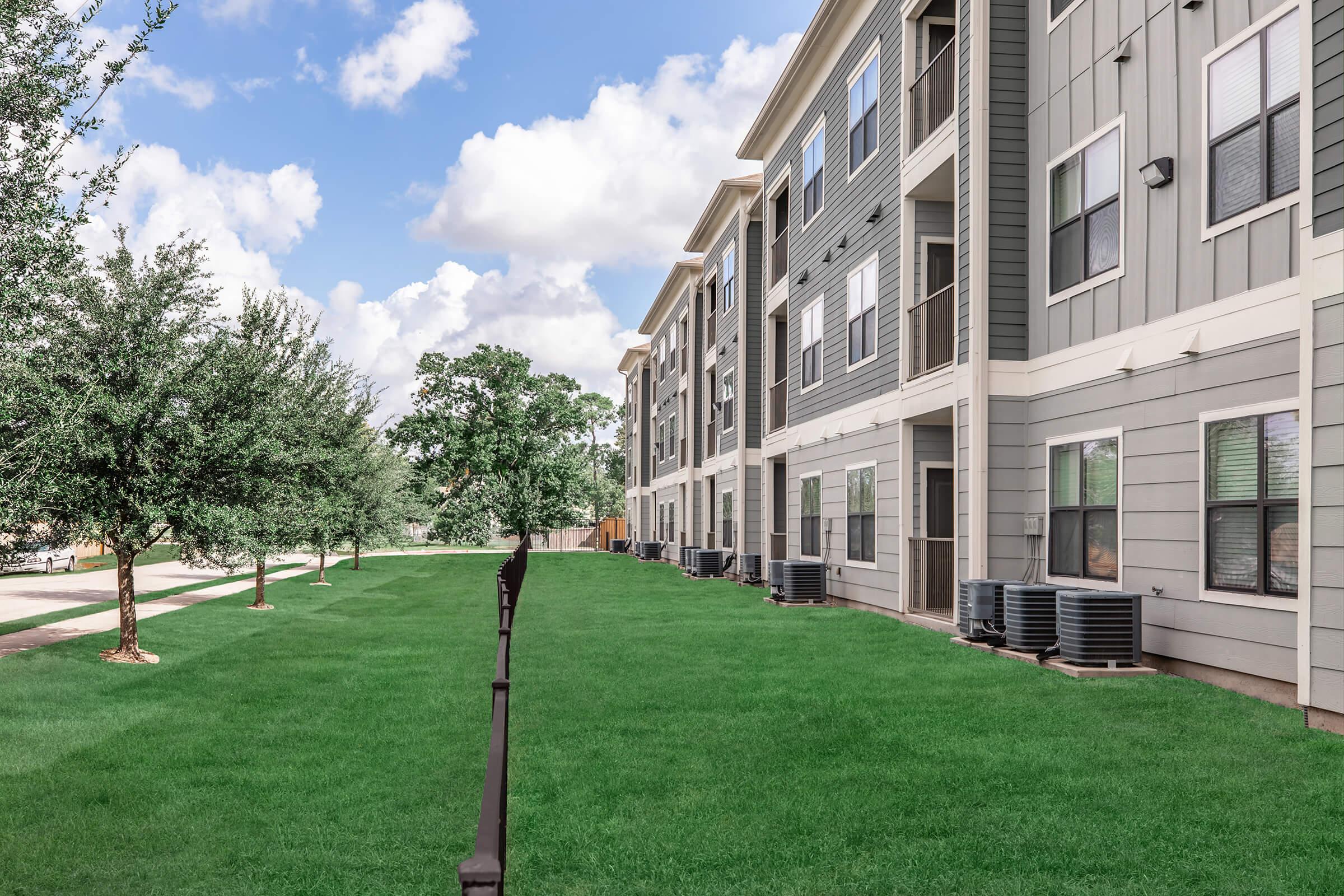 A well-maintained apartment complex features modern buildings with large windows and greenery. The image shows a lush, green lawn with small trees lining the walkway, providing a pleasant outdoor environment against a backdrop of fluffy white clouds in a blue sky.