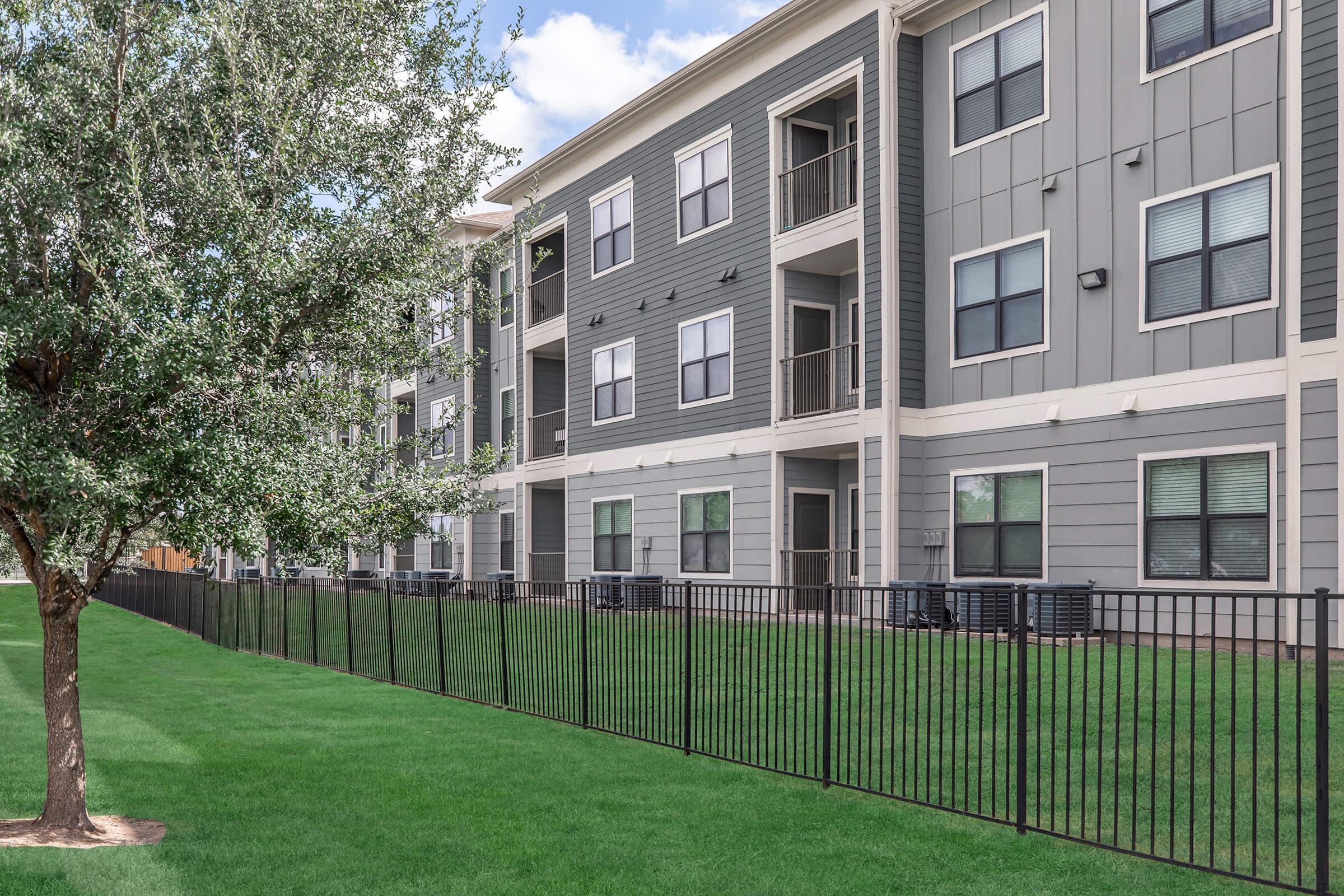 View of a modern apartment building with multiple levels, featuring a gray exterior. The ground is green and well-maintained, with a black fence lining the property. Trees are visible in the foreground, providing a natural touch to the urban setting. Blue sky and fluffy clouds are in the background.