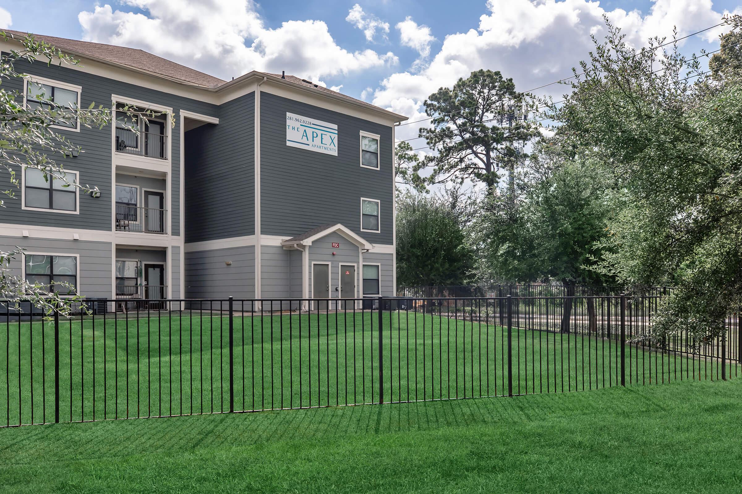 A multi-story apartment building with a gray exterior, featuring large windows and a sign that reads "APEX." In front, there is a well-maintained green lawn bordered by a black metal fence, with some trees visible in the background under a partly cloudy sky.