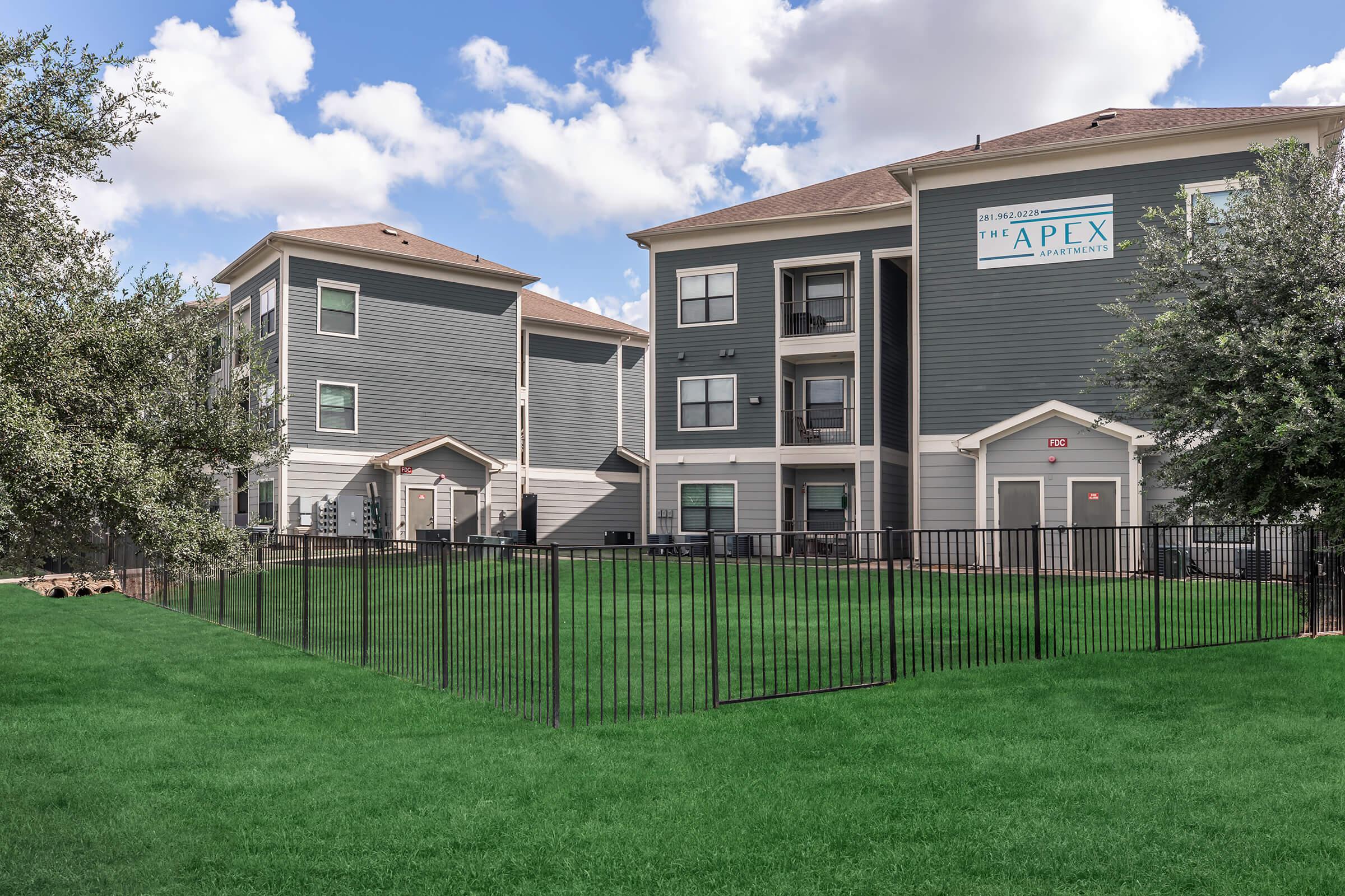 View of a modern apartment complex named "The Apex" featuring three buildings with a landscaped lawn area enclosed by a black metal fence. The sky is partly cloudy, enhancing the bright and inviting atmosphere of the property.