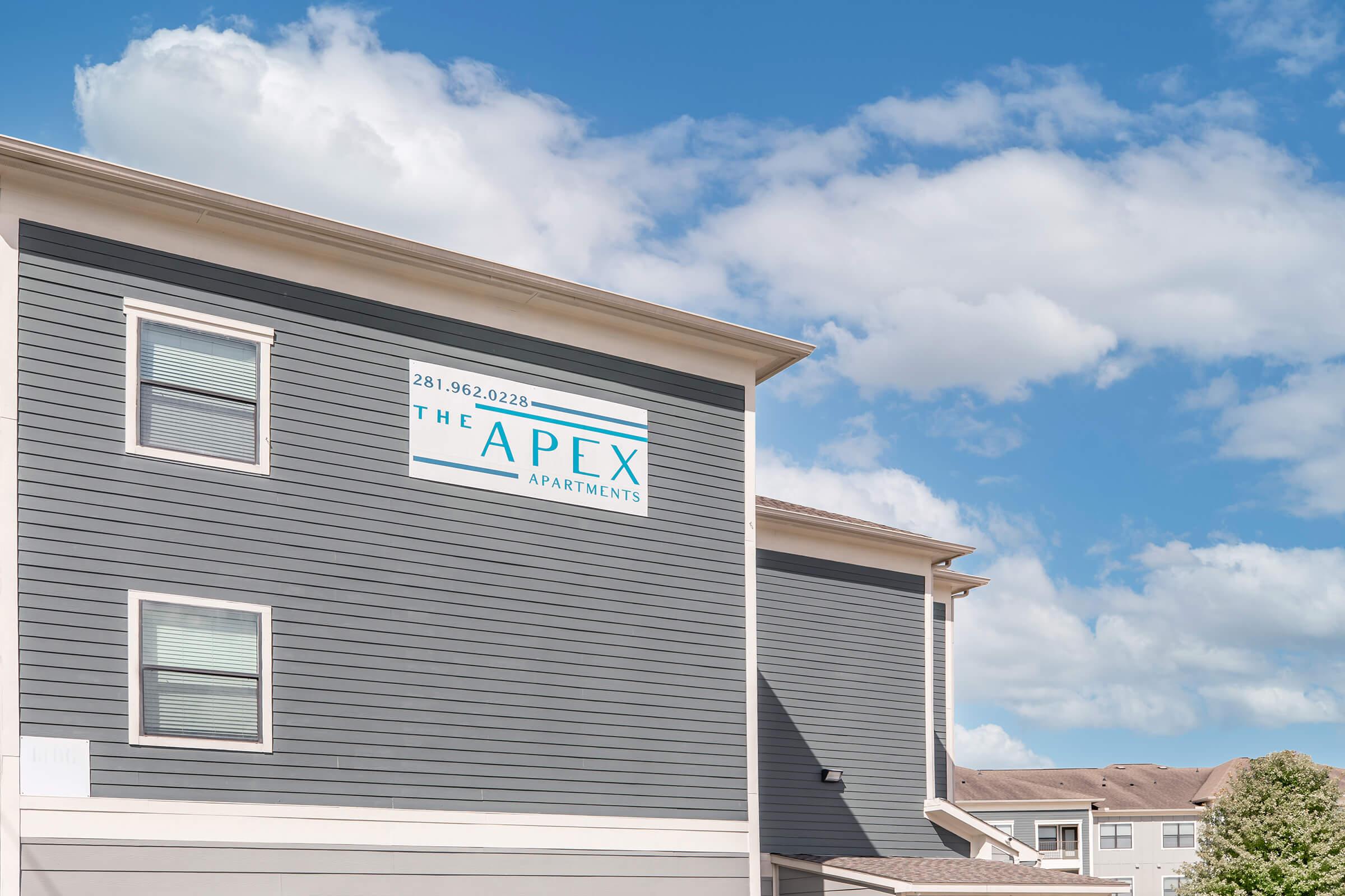 Signage on the side of a building for "The Apex Apartments," featuring the phone number 281-962-2028, set against a blue sky with white clouds. The building has a modern design with gray siding and several windows.